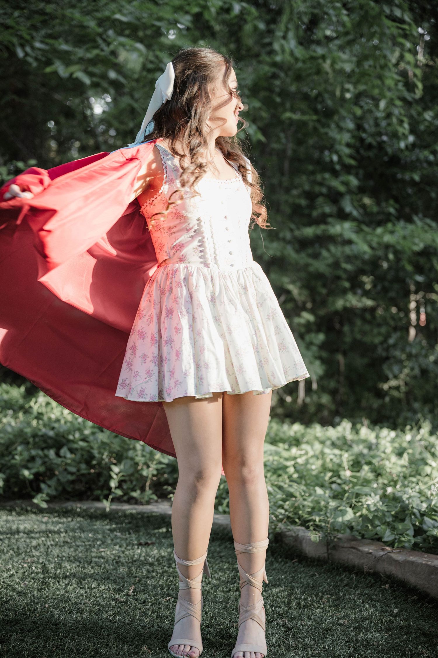 Woman in a white floral mini dress and lace-up sandals twirls a flowing red cape in a green garden.