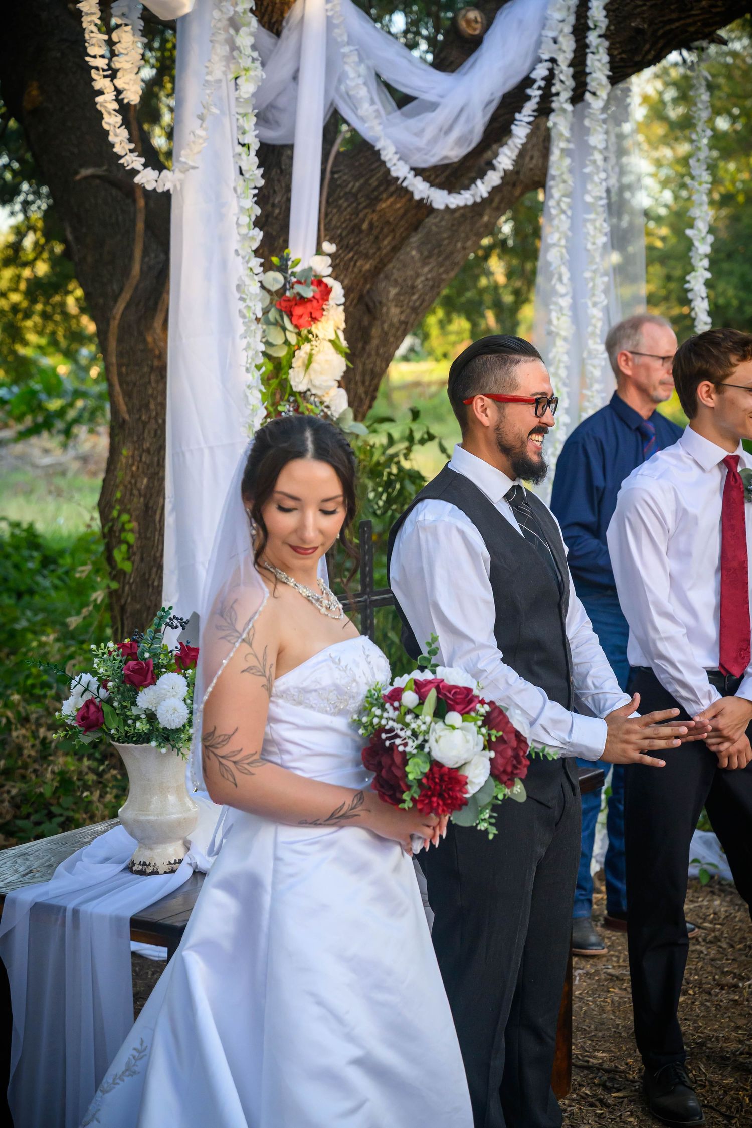 Bride and groom stand at outdoor wedding altar under white draped canopy, bride holding red and white bouquet.