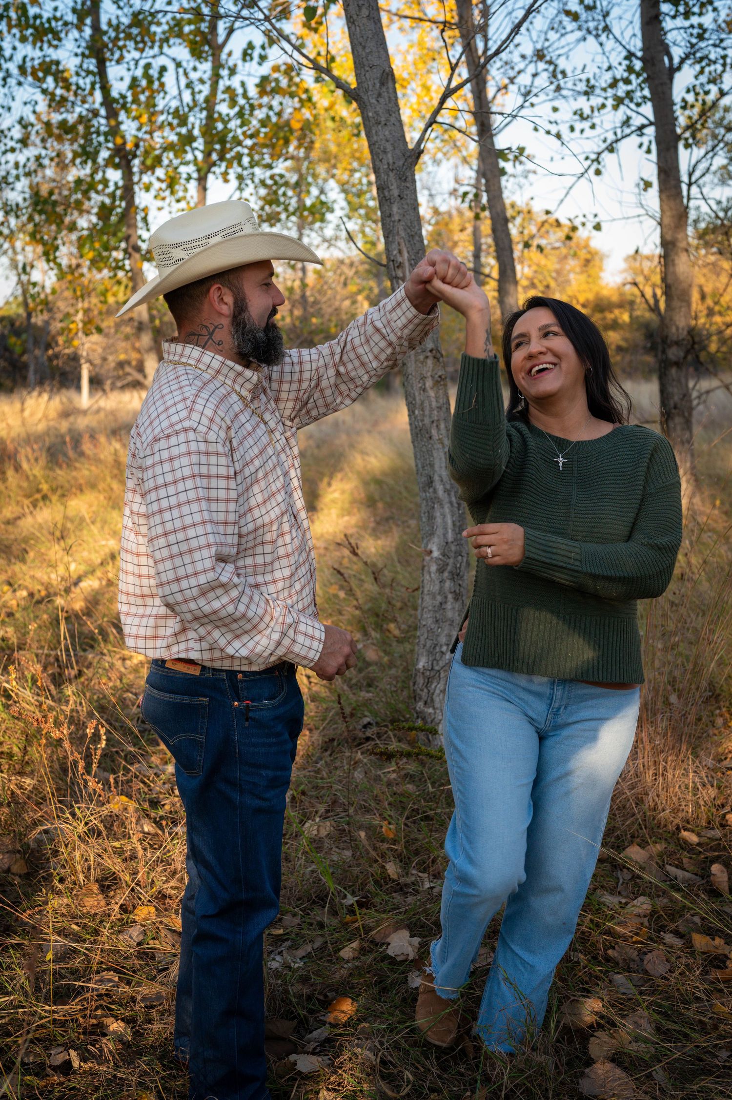 A couple shares a playful moment dancing together among autumn trees at sunset in casual western attire. Azle family photo session.