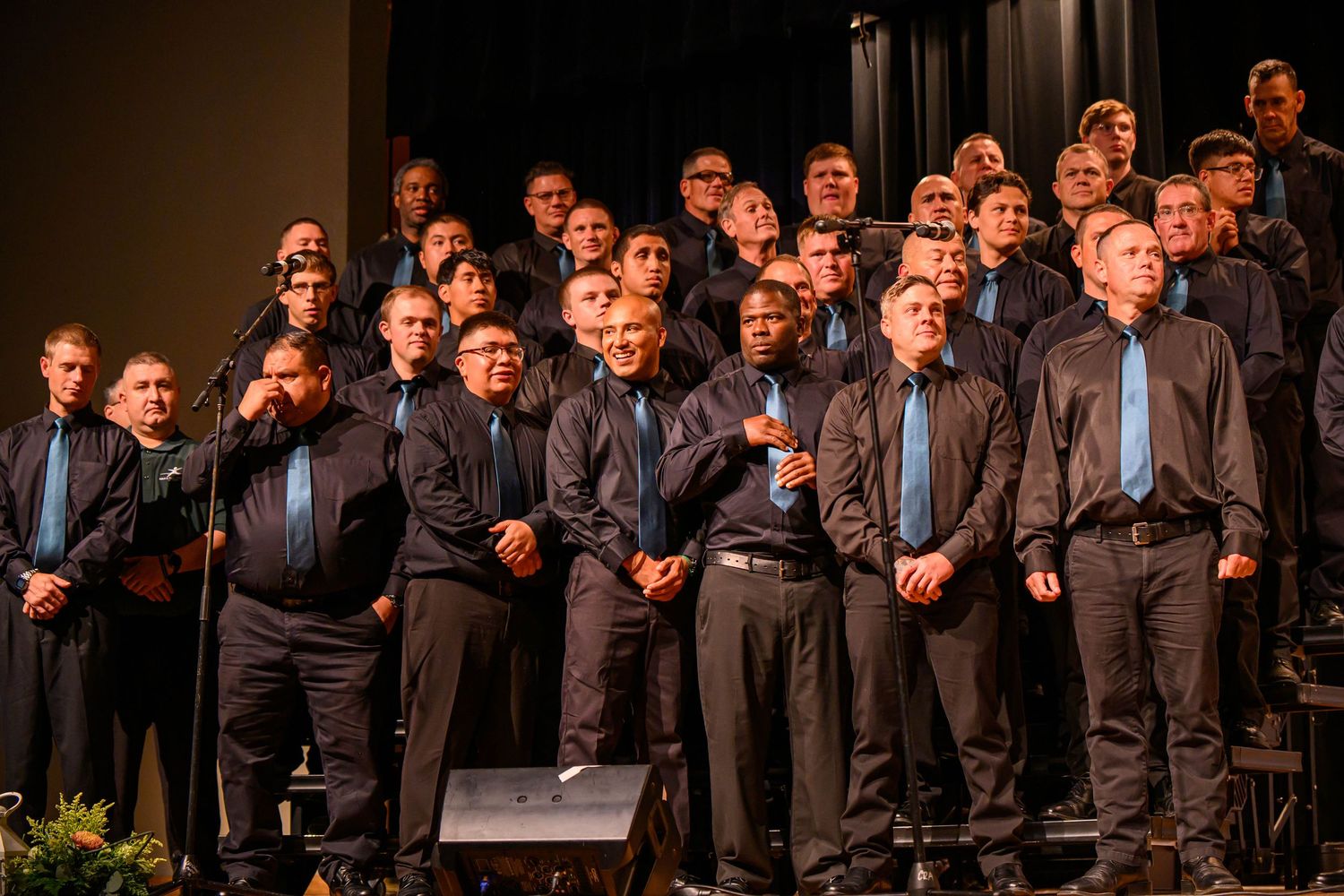 A large men's choir in dark shirts and blue ties stands in tiered rows on stage, singing during a live choral performance.