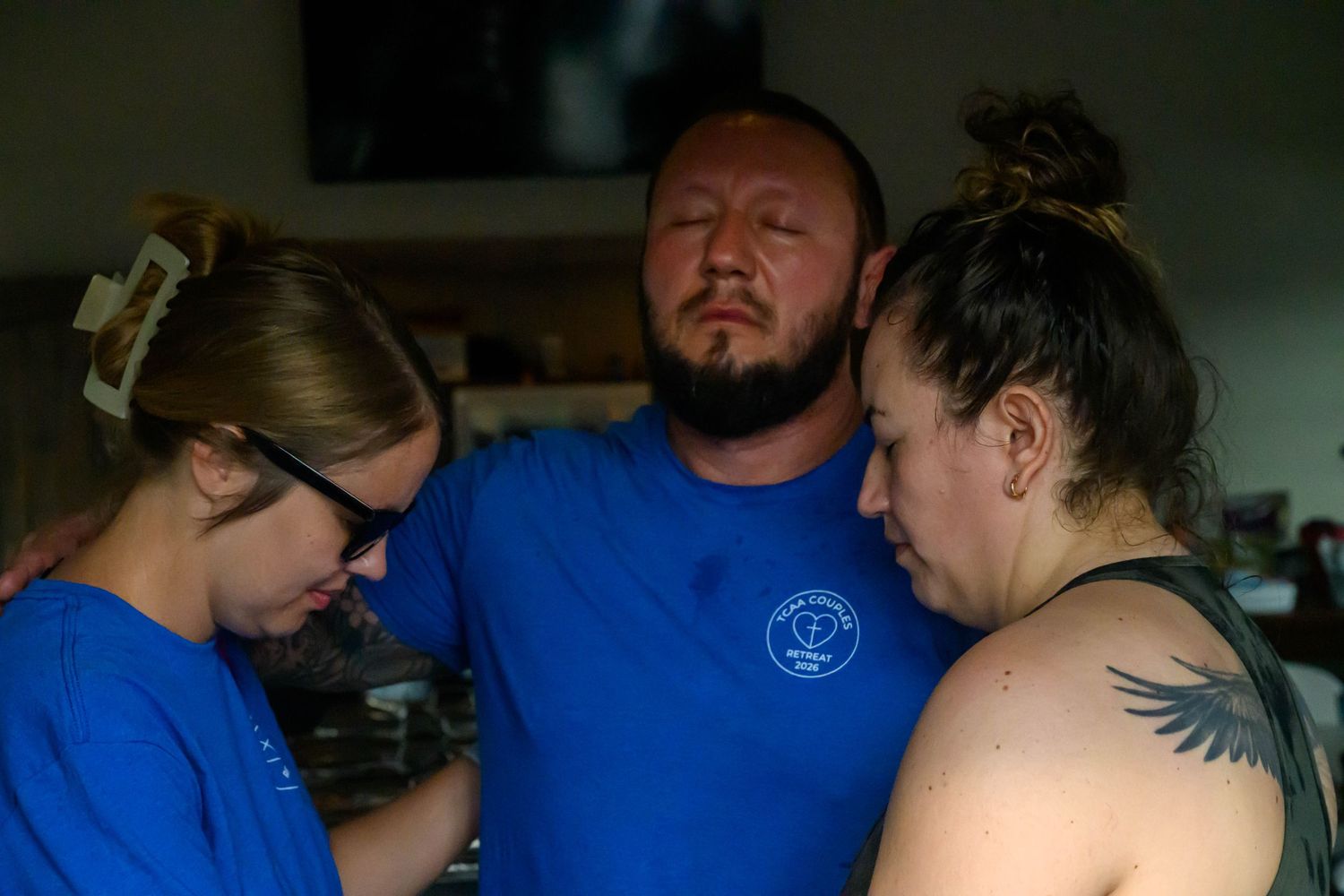Three people in blue shirts stand close together with heads bowed in prayer or quiet reflection inside a softly lit room.