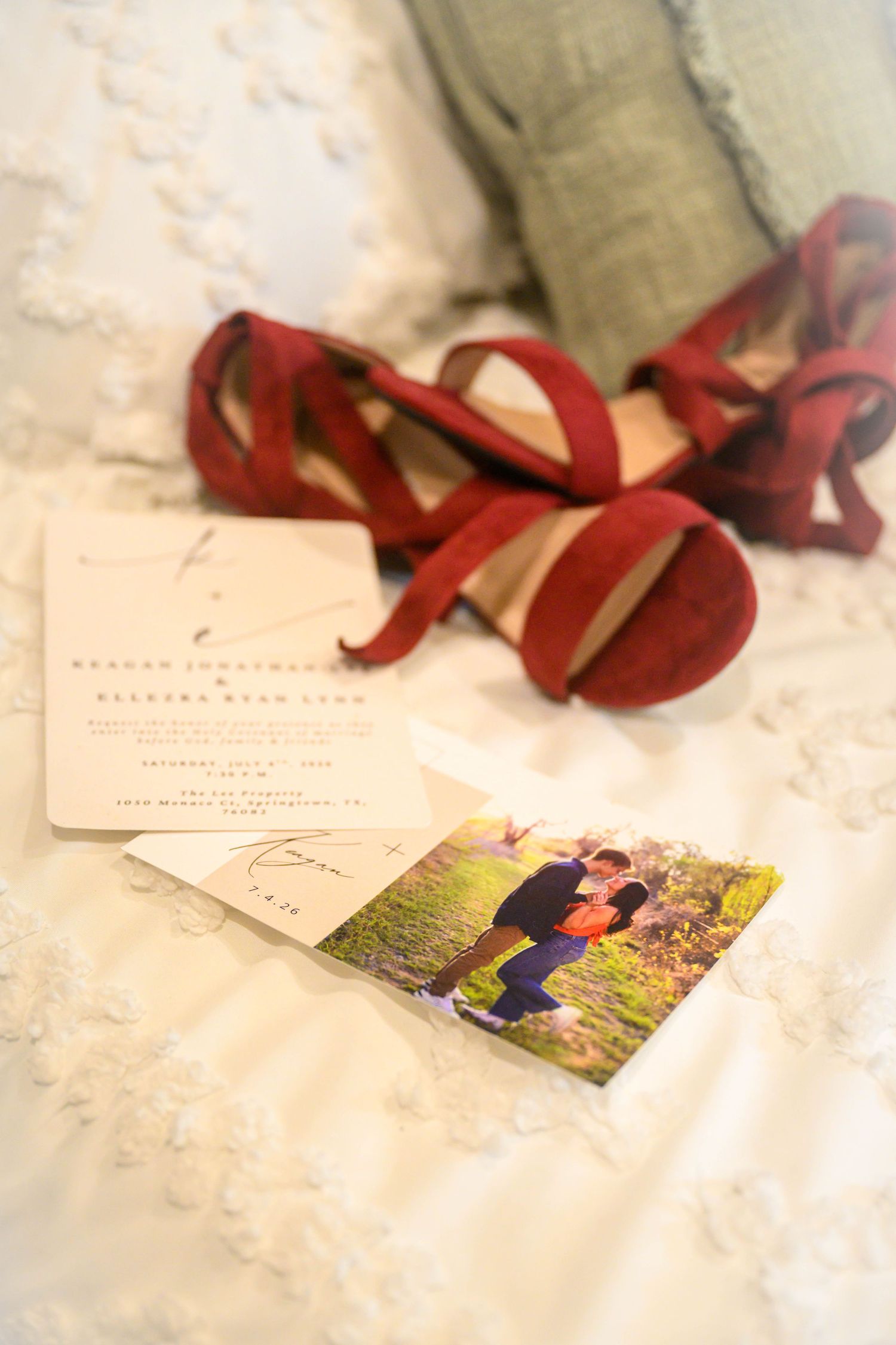 Red strappy wedding heels beside invitation cards and couple photo on white textured bedding.