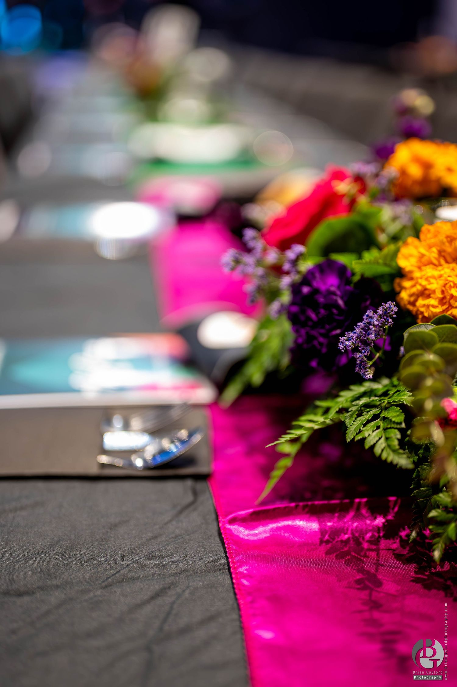 Vibrant pink, purple, and orange flowers arranged on a dark surface with green foliage create a striking contrast. Event Photograph.