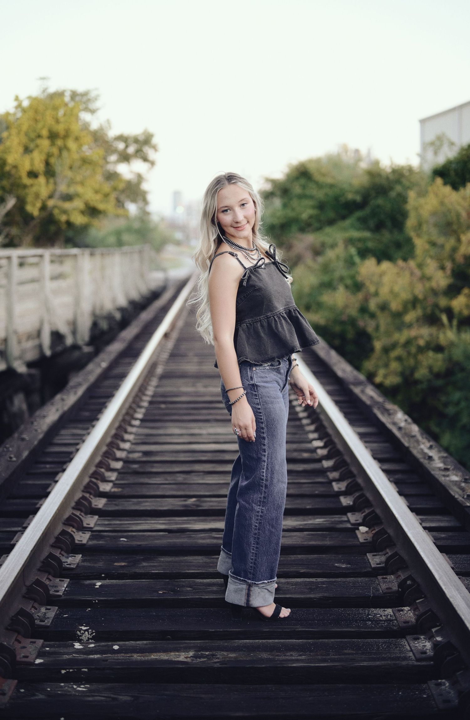 A child in denim overalls stands on wooden railroad tracks surrounded by greenery on a sunny day. Azle senior at the fort worth stockyards.