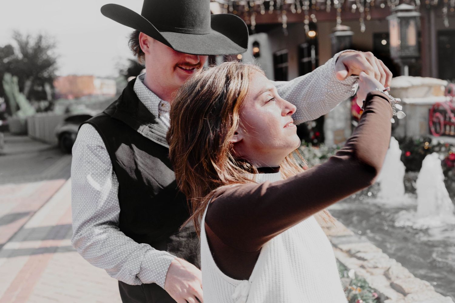 A man in a black cowboy hat and vest dances outdoors with a smiling woman in a white top near a sunlit fountain.