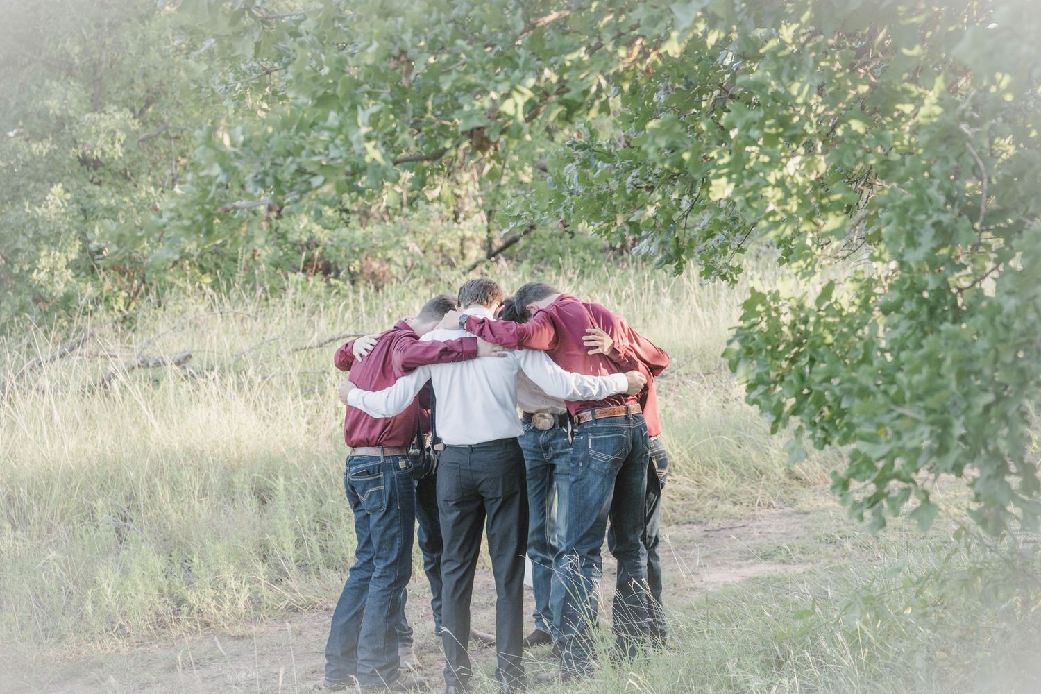 Group of people in maroon shirts and jeans standing in a close huddle with arms around each other on a dirt path beside green trees.