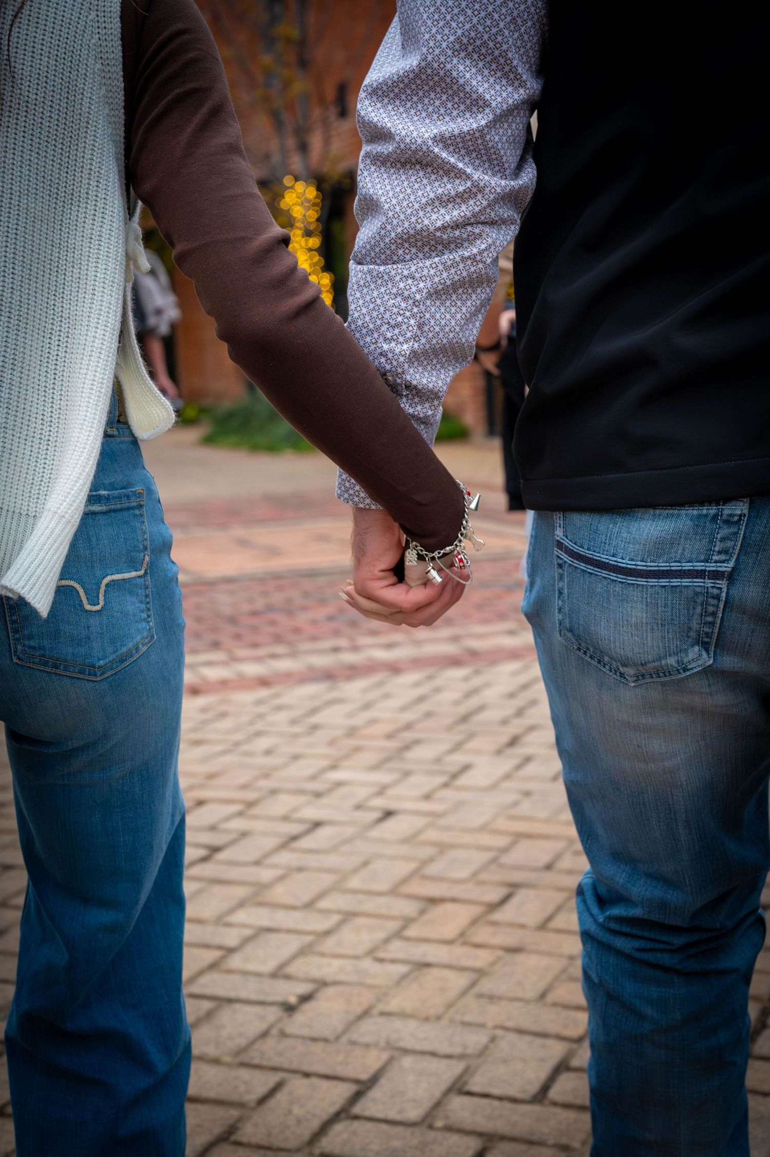 Two people in jeans holding hands while standing on a brick-paved walkway, with a bracelet visible on one wrist.