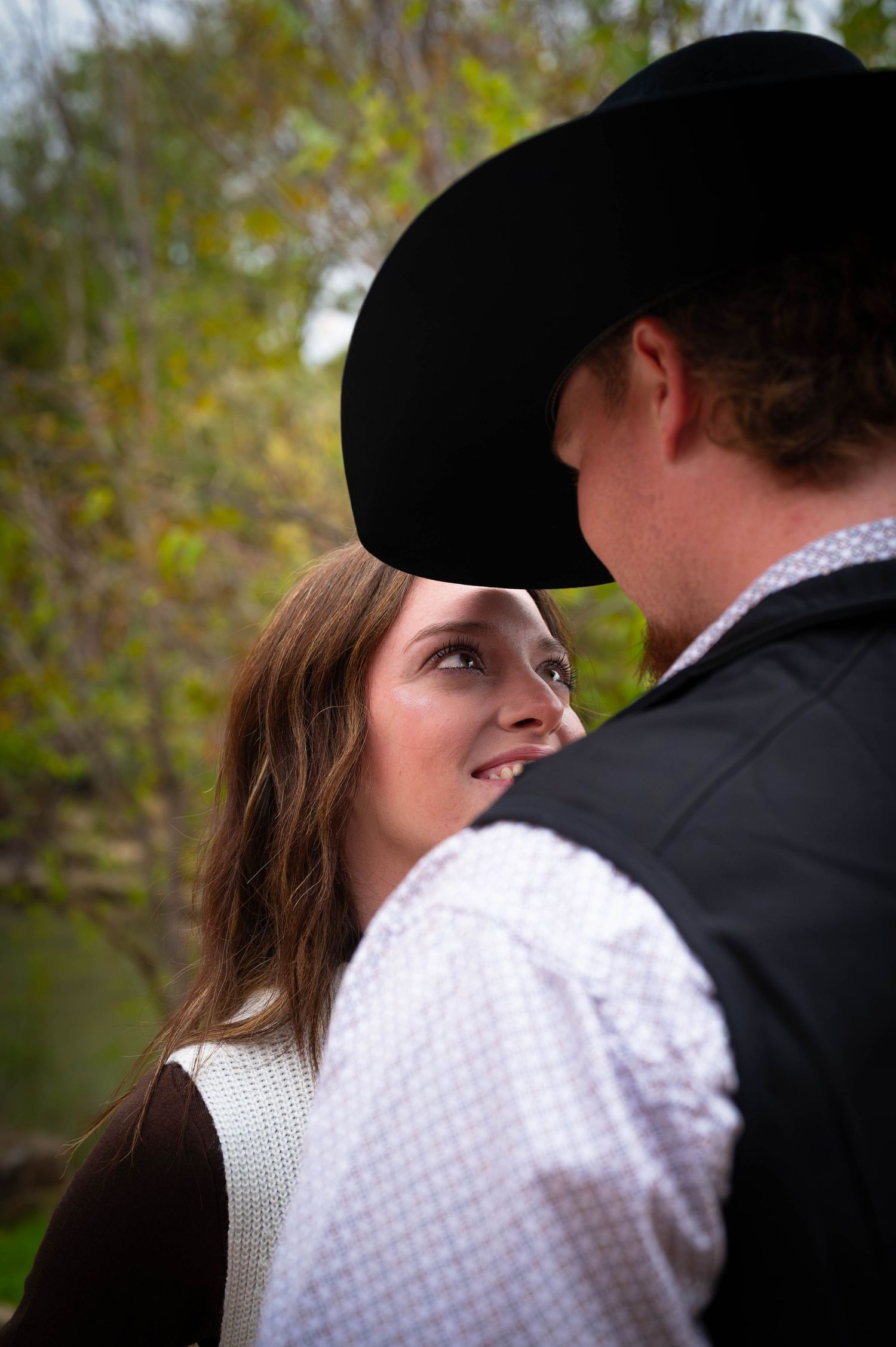 A smiling woman with long brown hair gazes up at a man in a black cowboy hat and vest in an outdoor autumn setting.