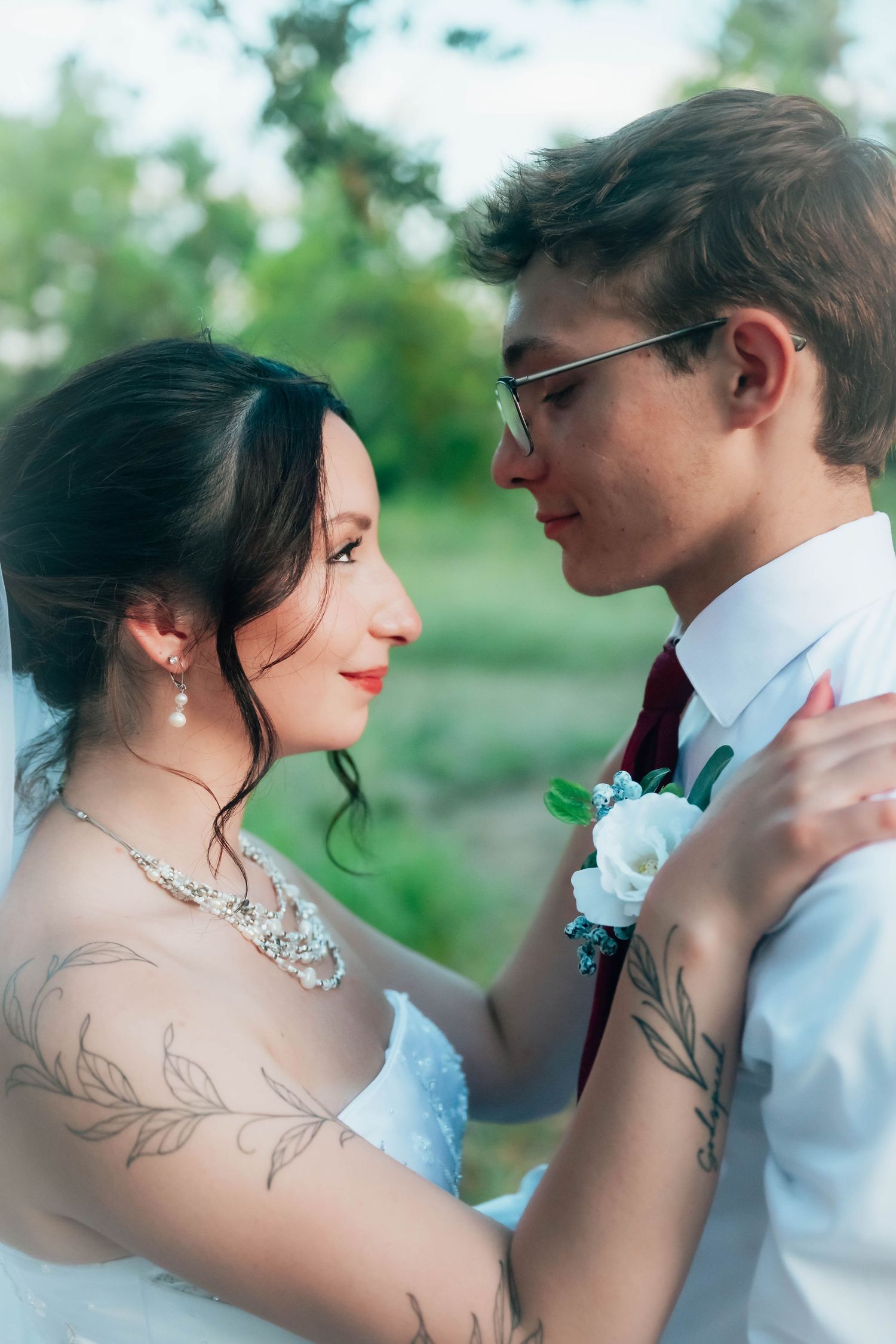 A bride and groom share an intimate moment outdoors, with the tattooed bride in a white dress and groom in a white shirt with boutonniere.