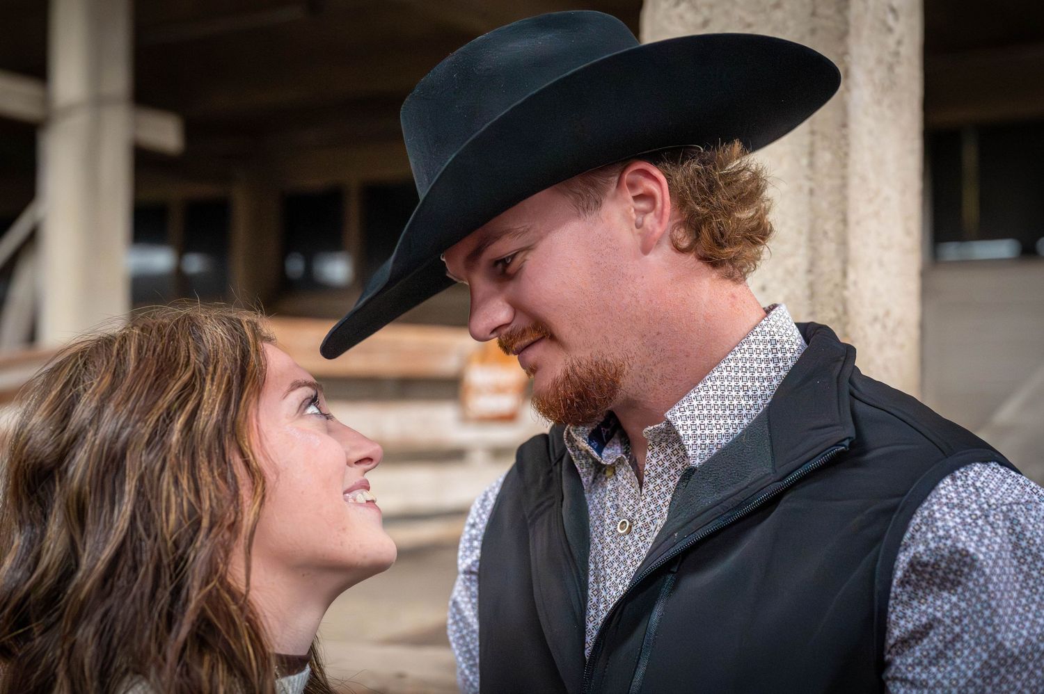 Smiling couple gazing at each other, a bearded man in a black cowboy hat and vest leaning toward a woman with long brown hair.