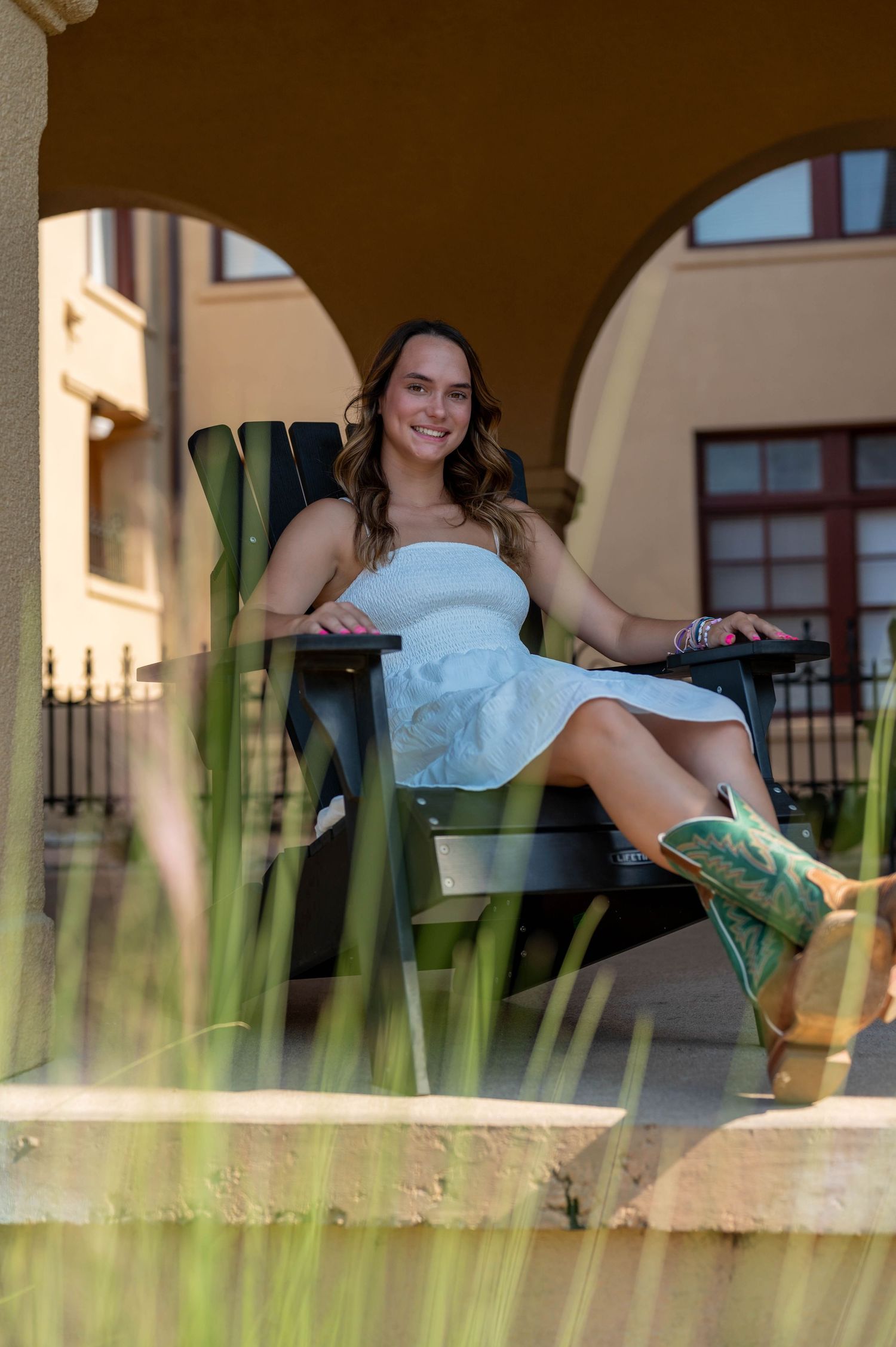 Smiling young woman in a white sundress and green cowboy boots relaxes in a dark Adirondack chair under sunlit stone archways.
