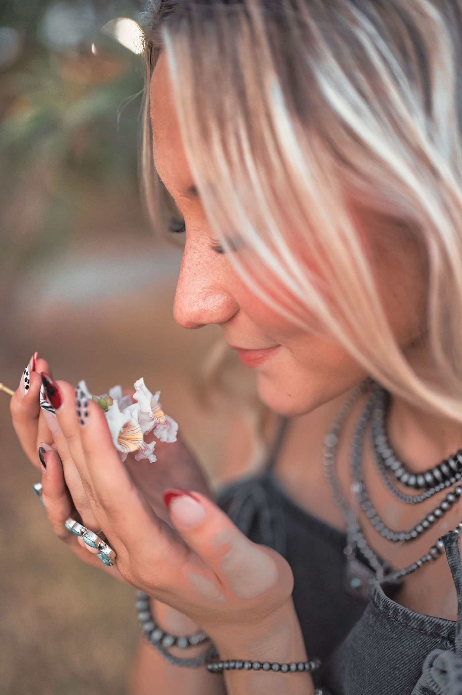 Blonde woman with freckles smiles while smelling a small white flower, wearing layered beaded necklaces, a denim top and patterned nails.