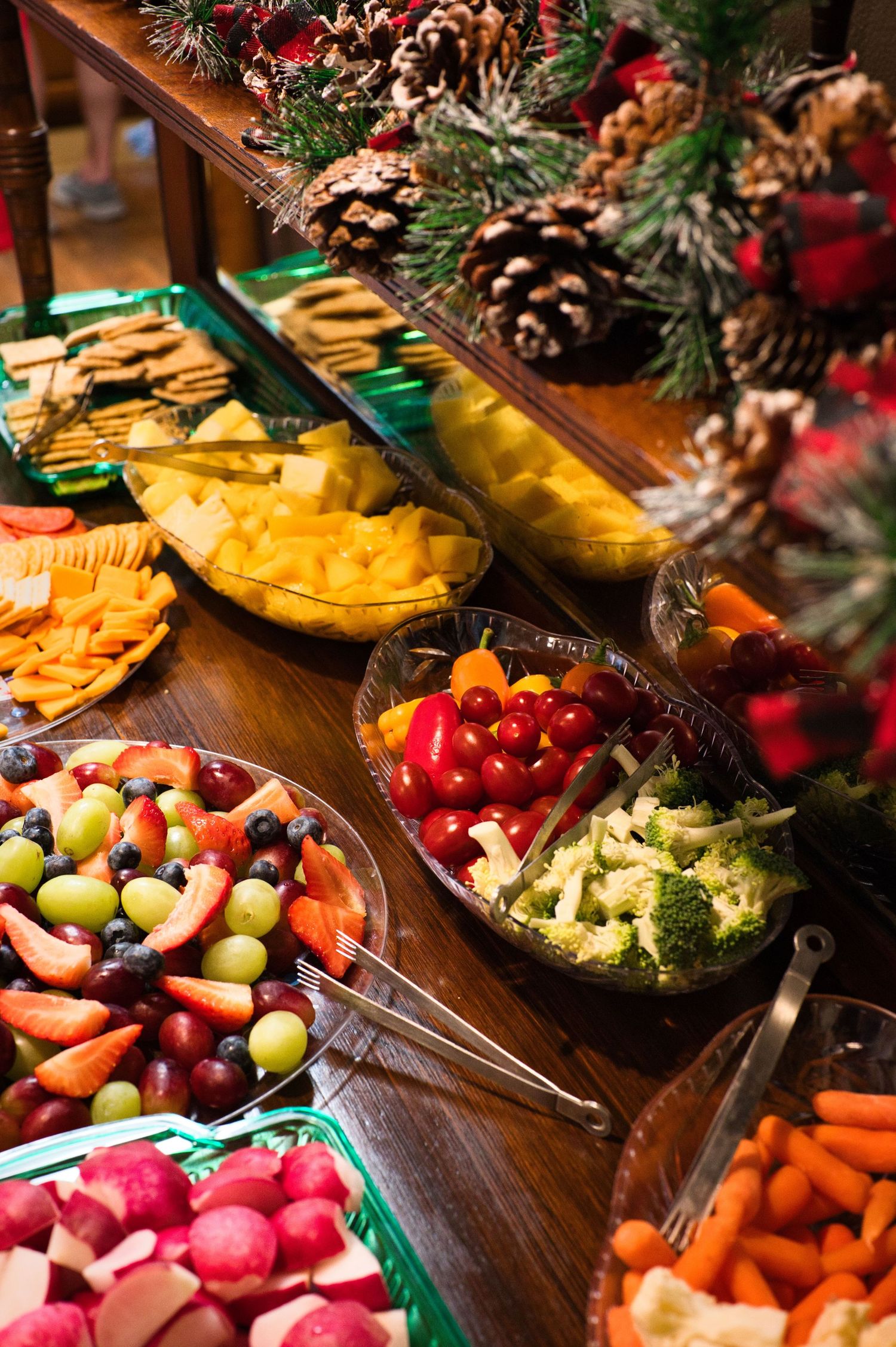 Festive holiday buffet table with fresh fruit, veggie and cheese platters, crackers, and pinecone and evergreen decorations.