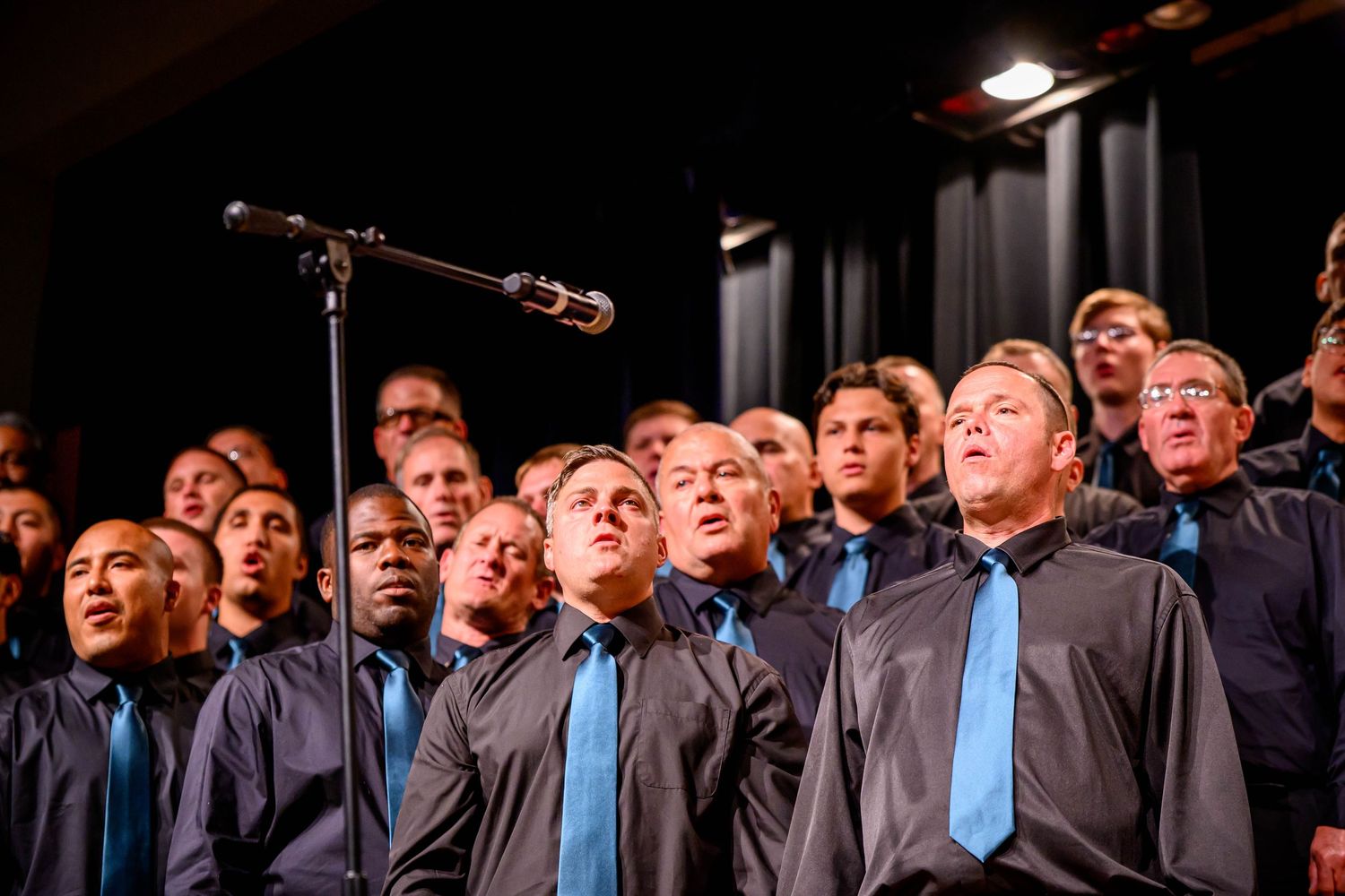 Men's chorus in matching gray shirts and blue ties singing on stage beside a microphone during a live choral performance.