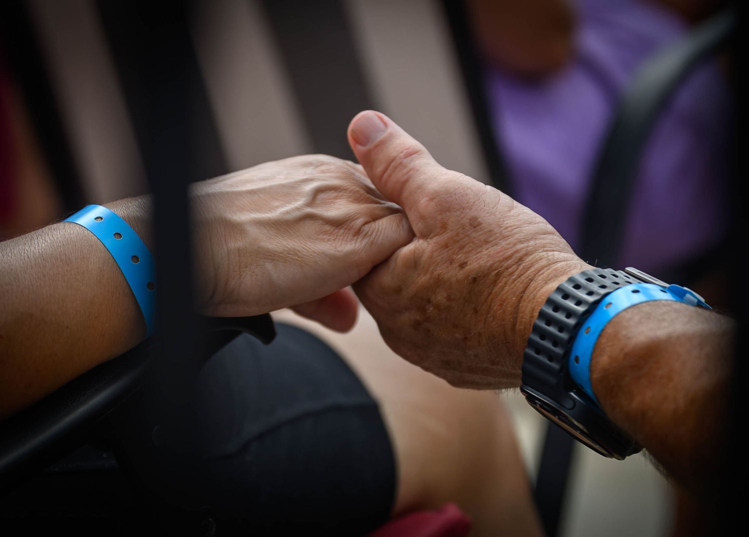 Two elderly hands clasped together in comfort, each wearing blue wristbands, with one wrist also wearing a black sport watch.