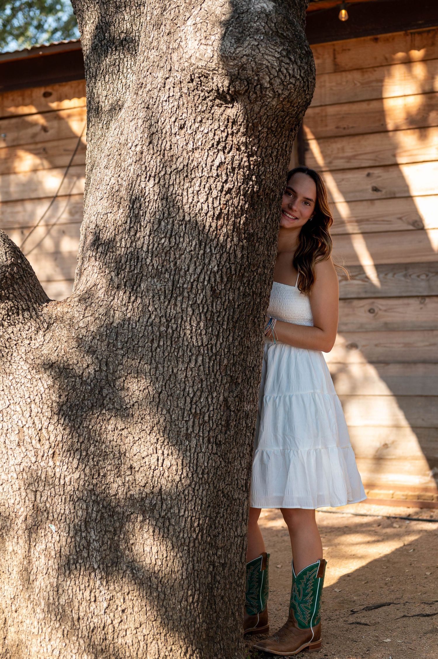 Smiling young woman in a white sundress and green cowboy boots peeks from behind a large tree trunk in front of a rustic wooden building.