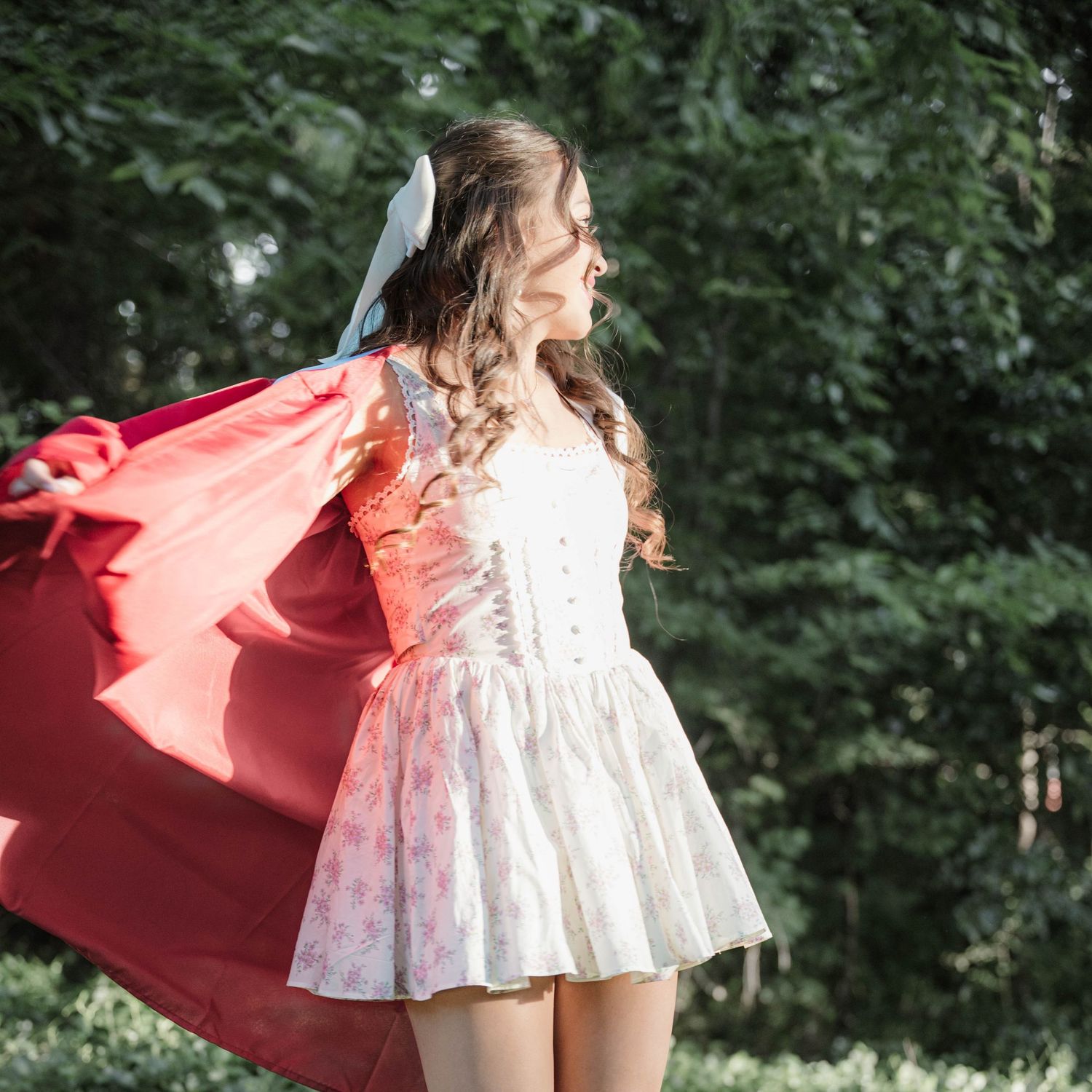 Young woman in white floral dress and flowing red cape twirling outdoors in a sunlit forest setting.