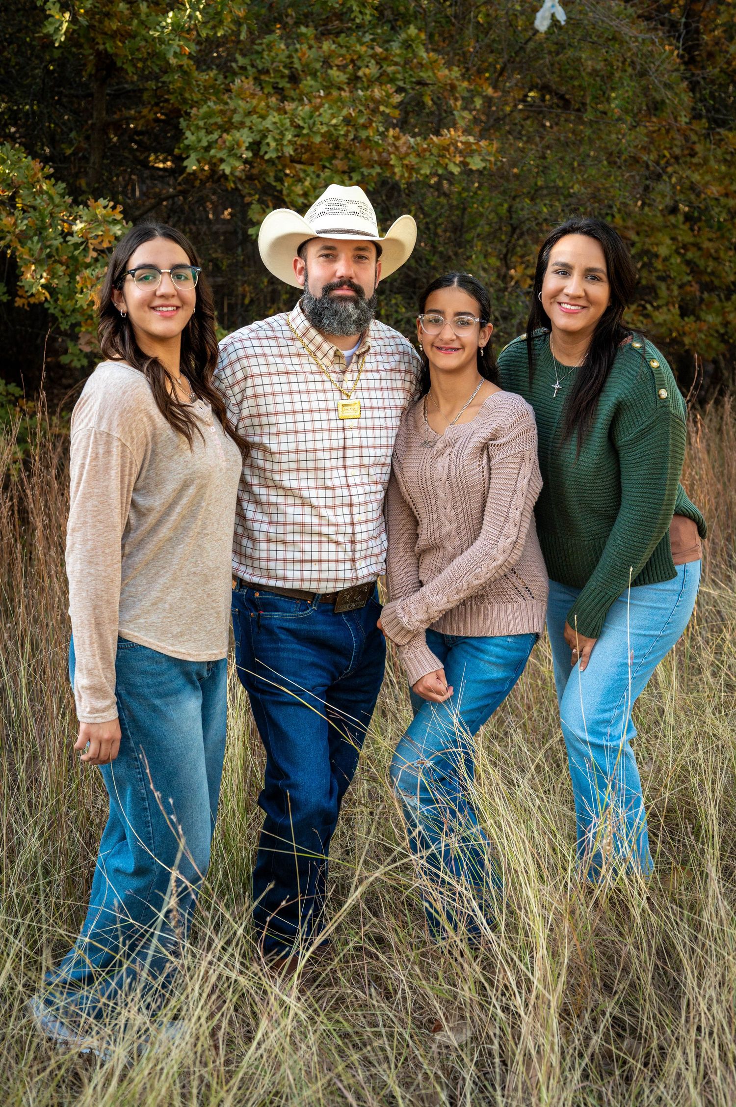 Family of four posing together in tall golden grass, a bearded man in a cowboy hat standing with three smiling women in casual outfits.