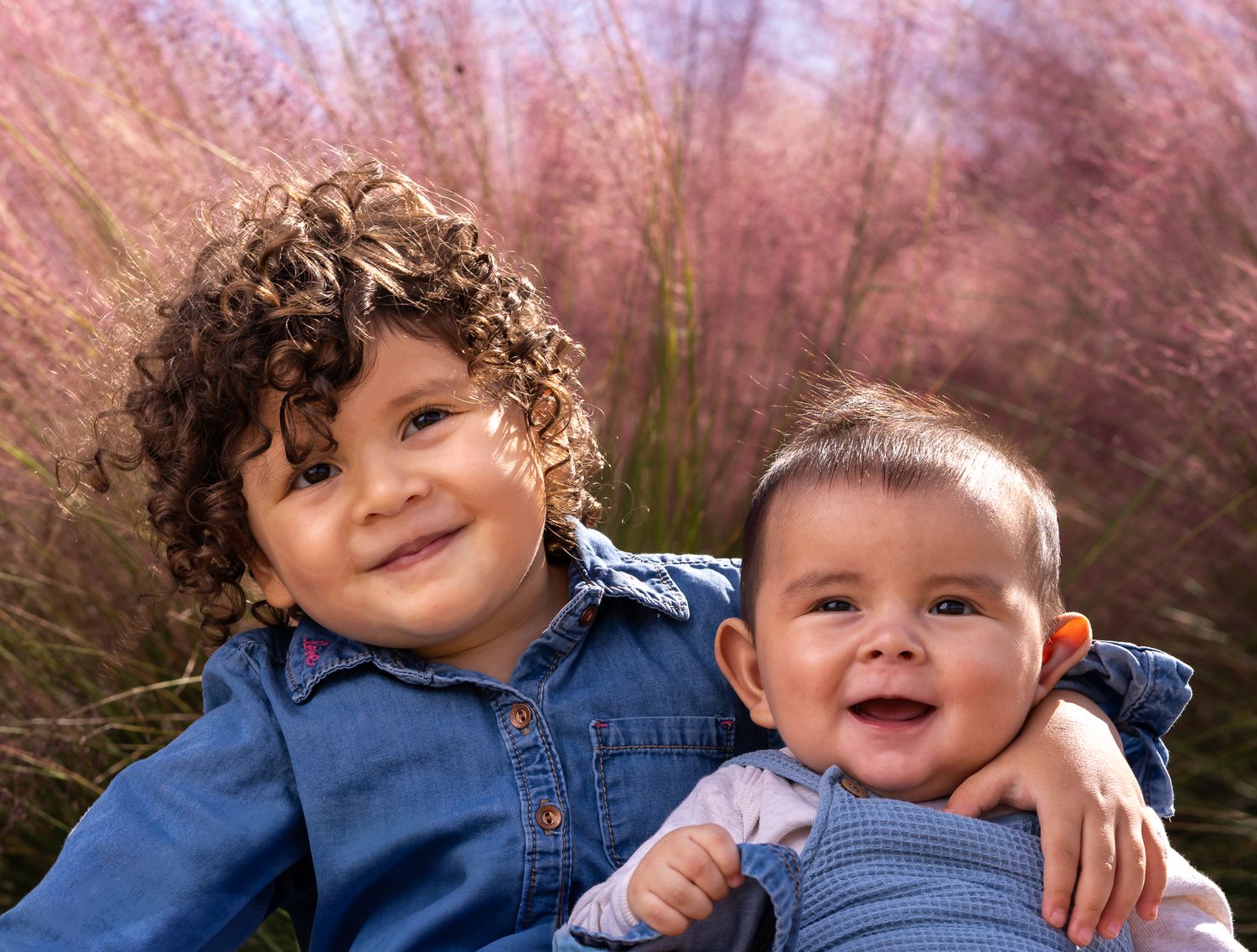 Two children in denim shirts smile while sitting together outdoors with pink blossoms in the background.