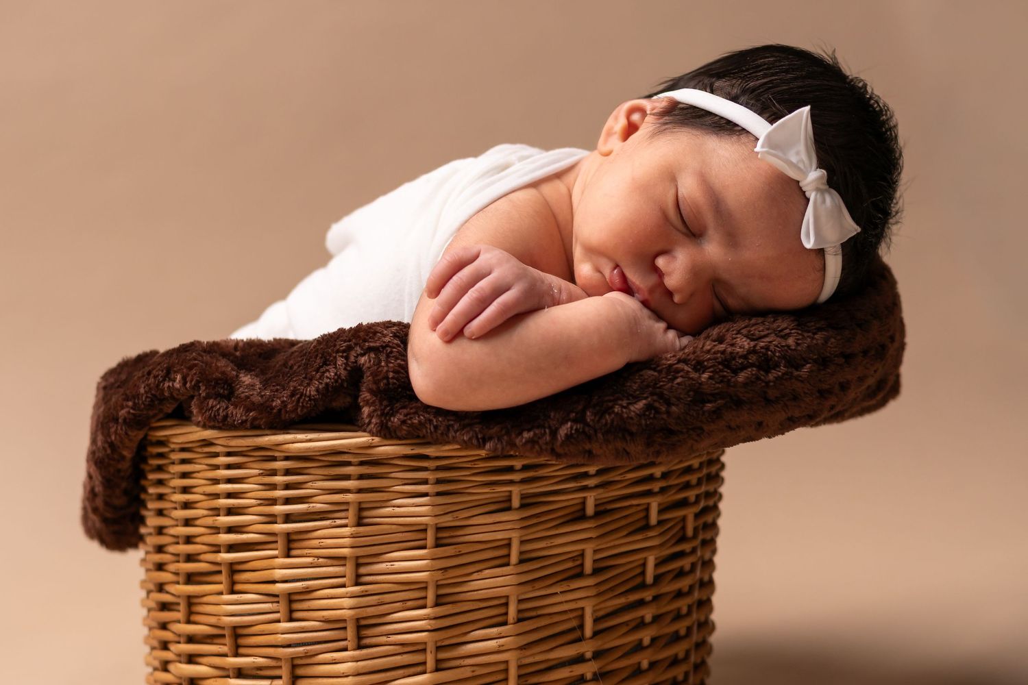Sleeping newborn in white wrap resting peacefully on brown fur blanket in wicker basket.