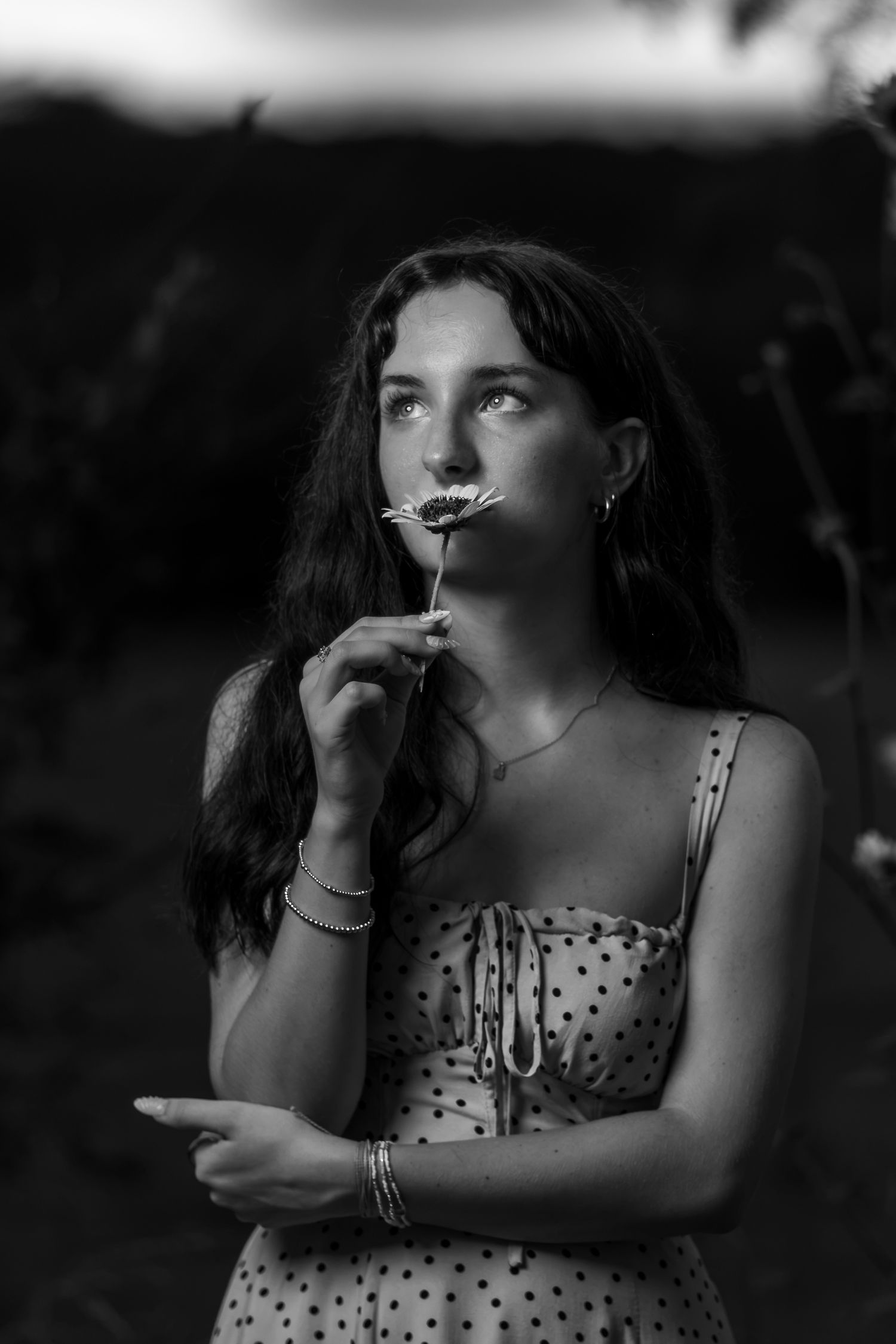 Black and white portrait of a young woman in a polka dot dress smelling a daisy flower outdoors.