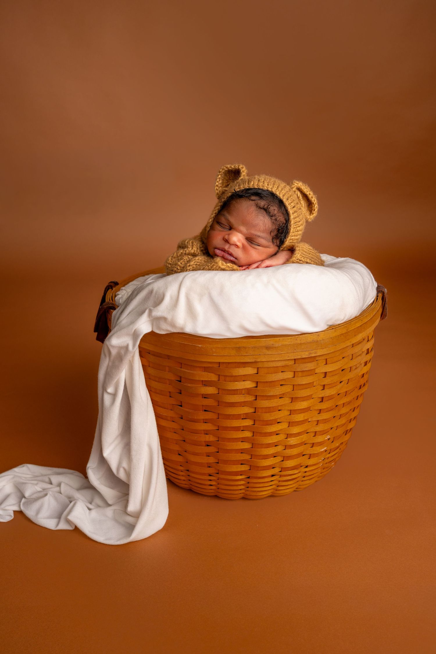 Newborn baby sleeping in a wicker basket wearing a knit bear bonnet, wrapped in white fabric on brown background.