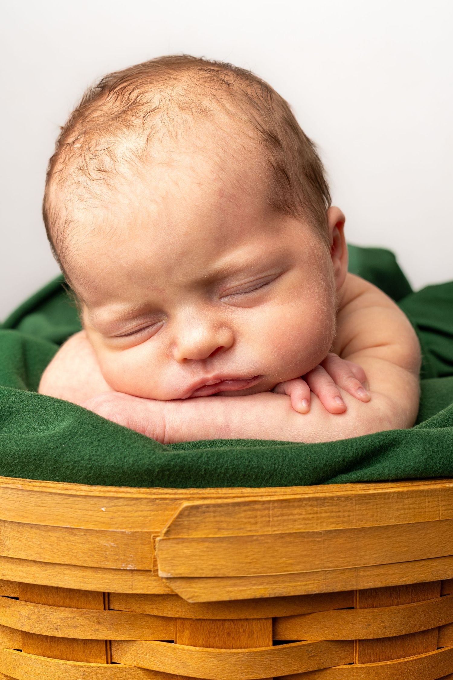 Sleeping newborn baby resting chin on hands inside a wooden wicker basket lined with green fabric.