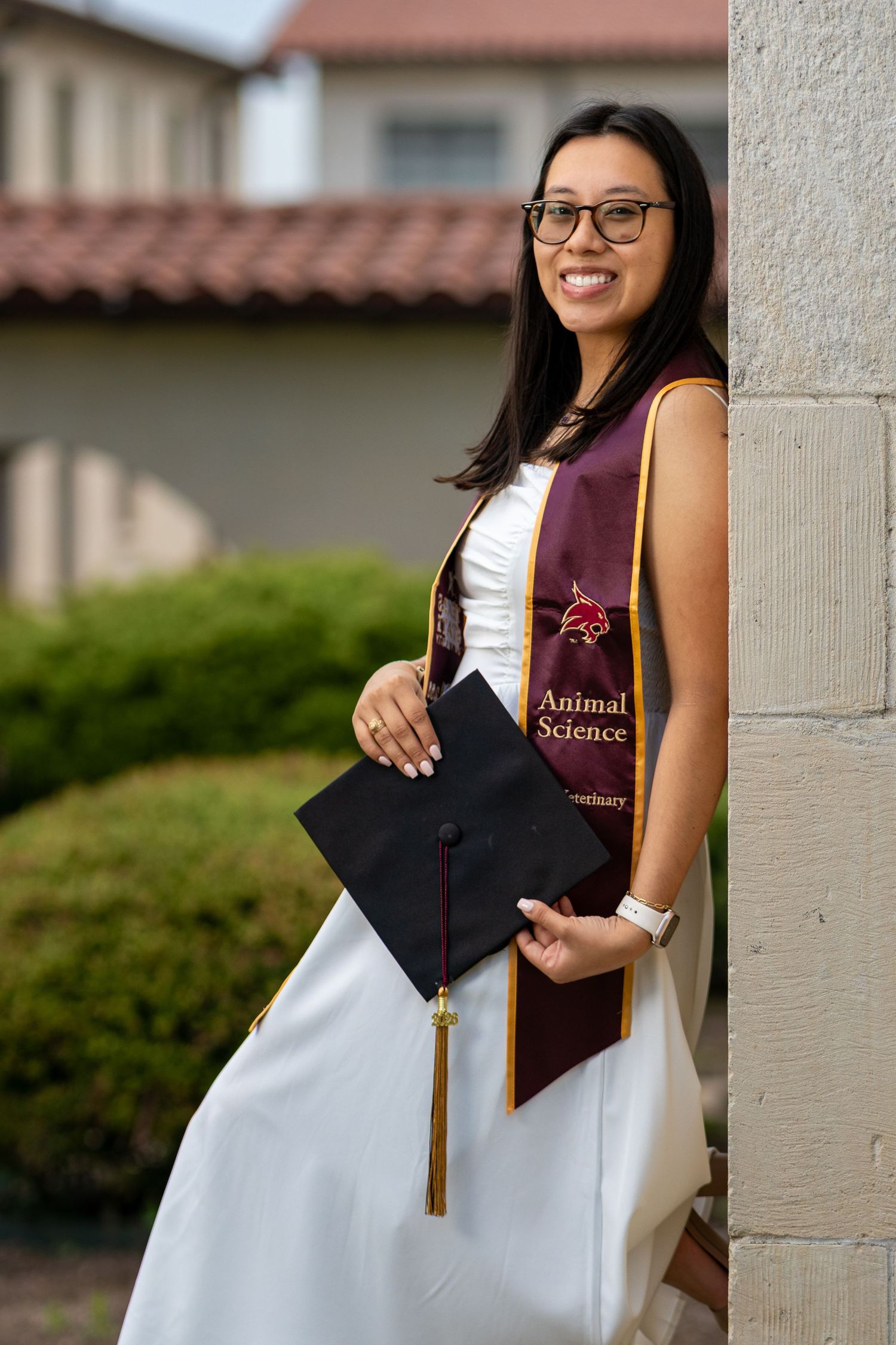 Smiling graduate in white dress holds black cap with Animal Science maroon stole against pillar.