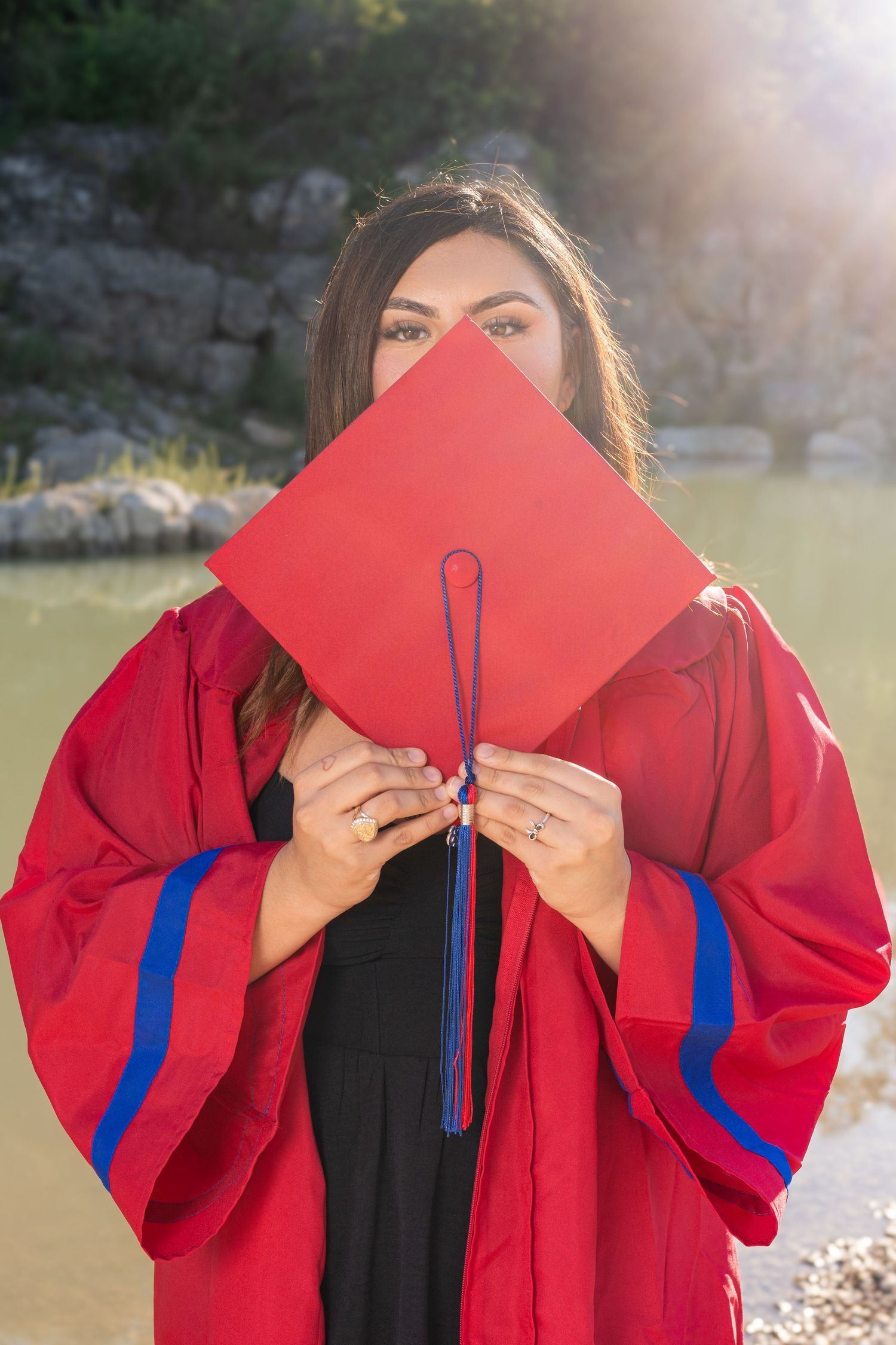 Graduate in red cap and gown holding mortarboard in front of face near sunlit river.