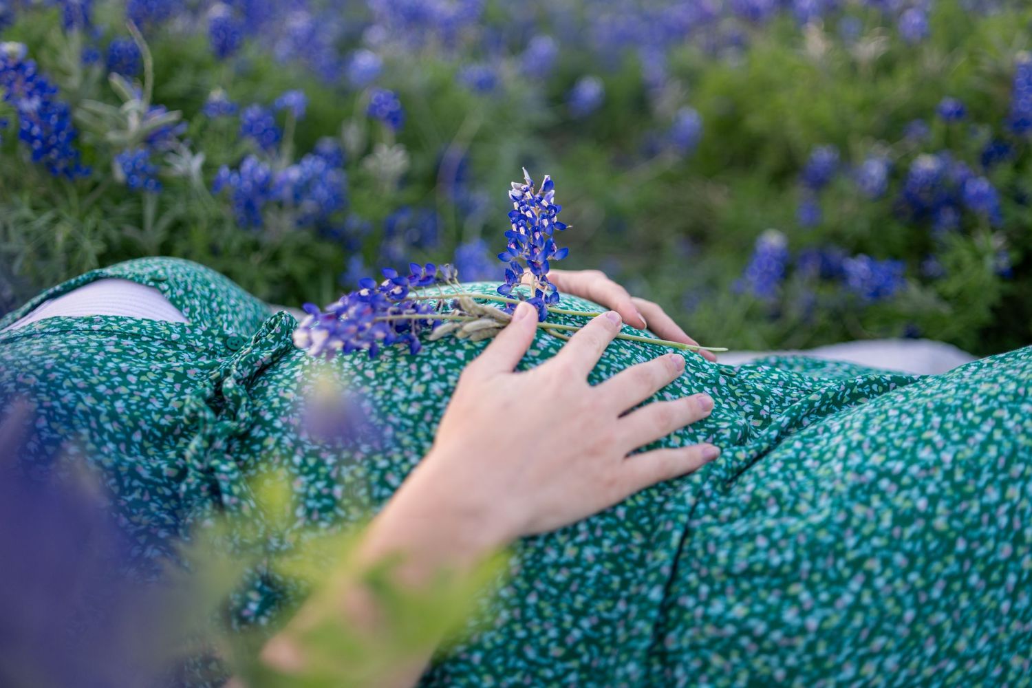 Pregnant woman in green floral dress holding bluebonnets while resting in a field of purple wildflowers.