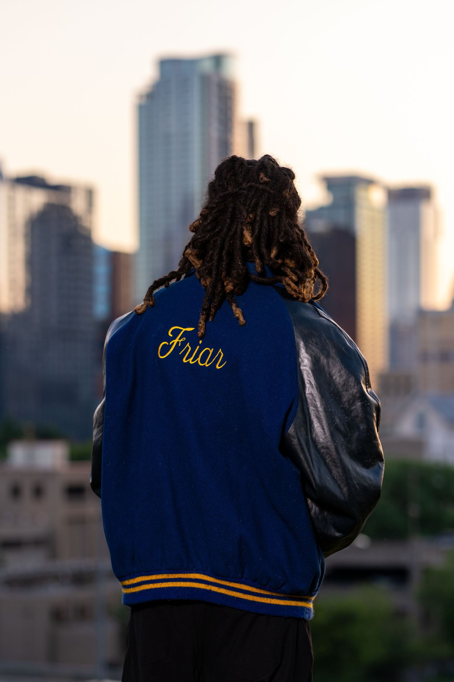 Person with dreadlocks wearing a blue Friar varsity jacket overlooking a city skyline at sunset.