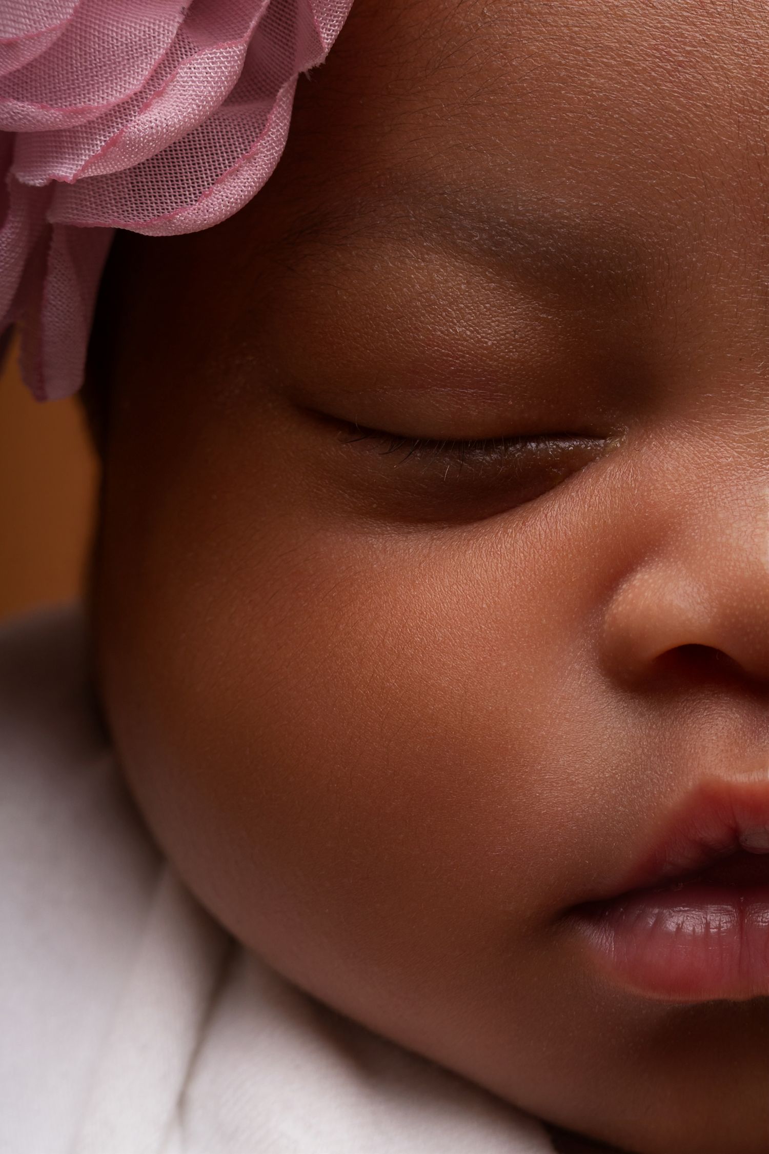 Close-up of a sleeping newborn baby with soft skin, closed eyes, and a pink flower headband resting against a white blanket.