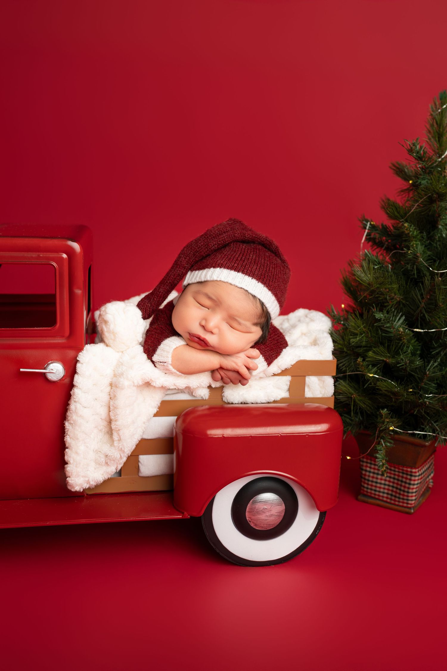 Sleeping newborn in a red knit Santa hat rests on a white blanket in a vintage red toy truck beside a small Christmas tree.