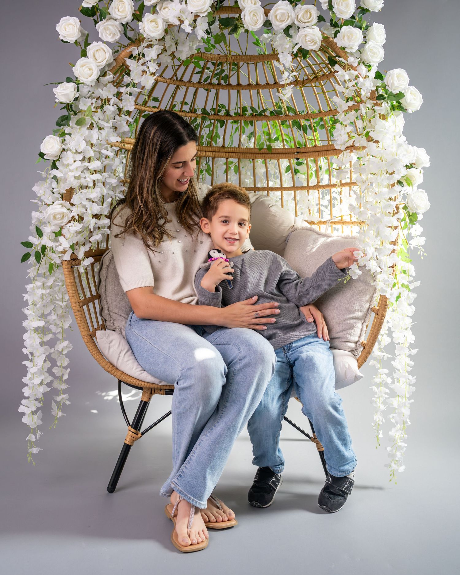 Mother and child share tender moments on a rattan chair decorated with white orchids against a grey backdrop.