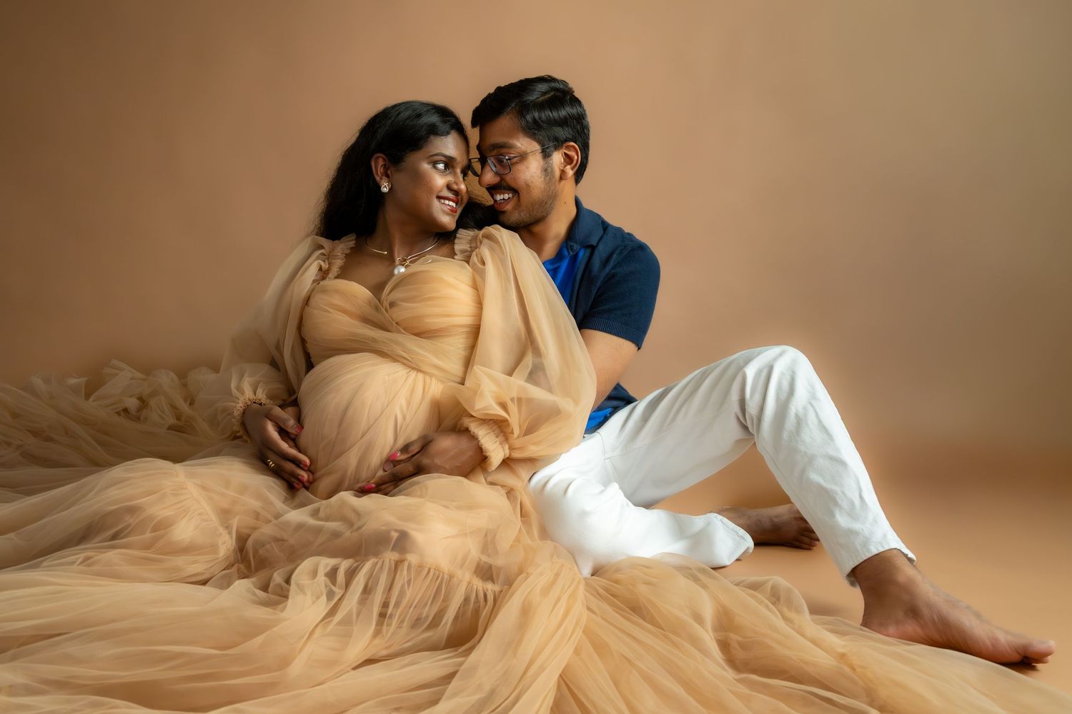 A pregnant woman in a flowing gold tulle gown sits with her partner in a warm studio maternity photoshoot.