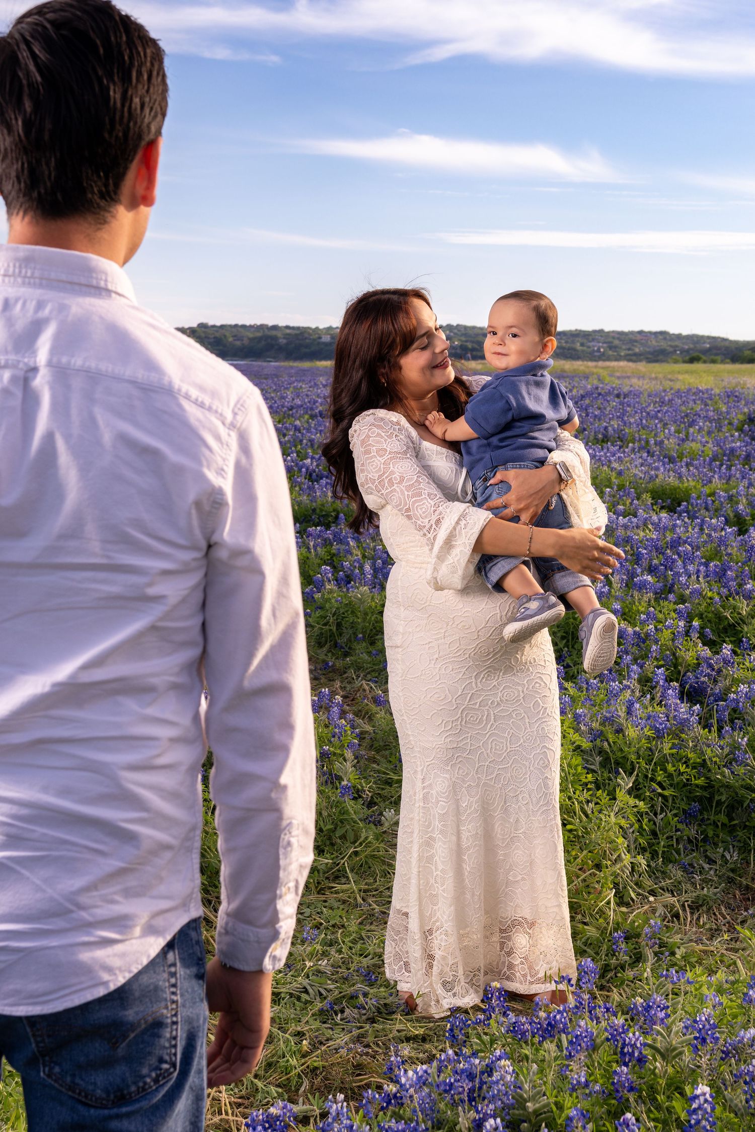 Family in bluebonnet field, woman in white lace dress holds baby boy, man in white shirt watches.
