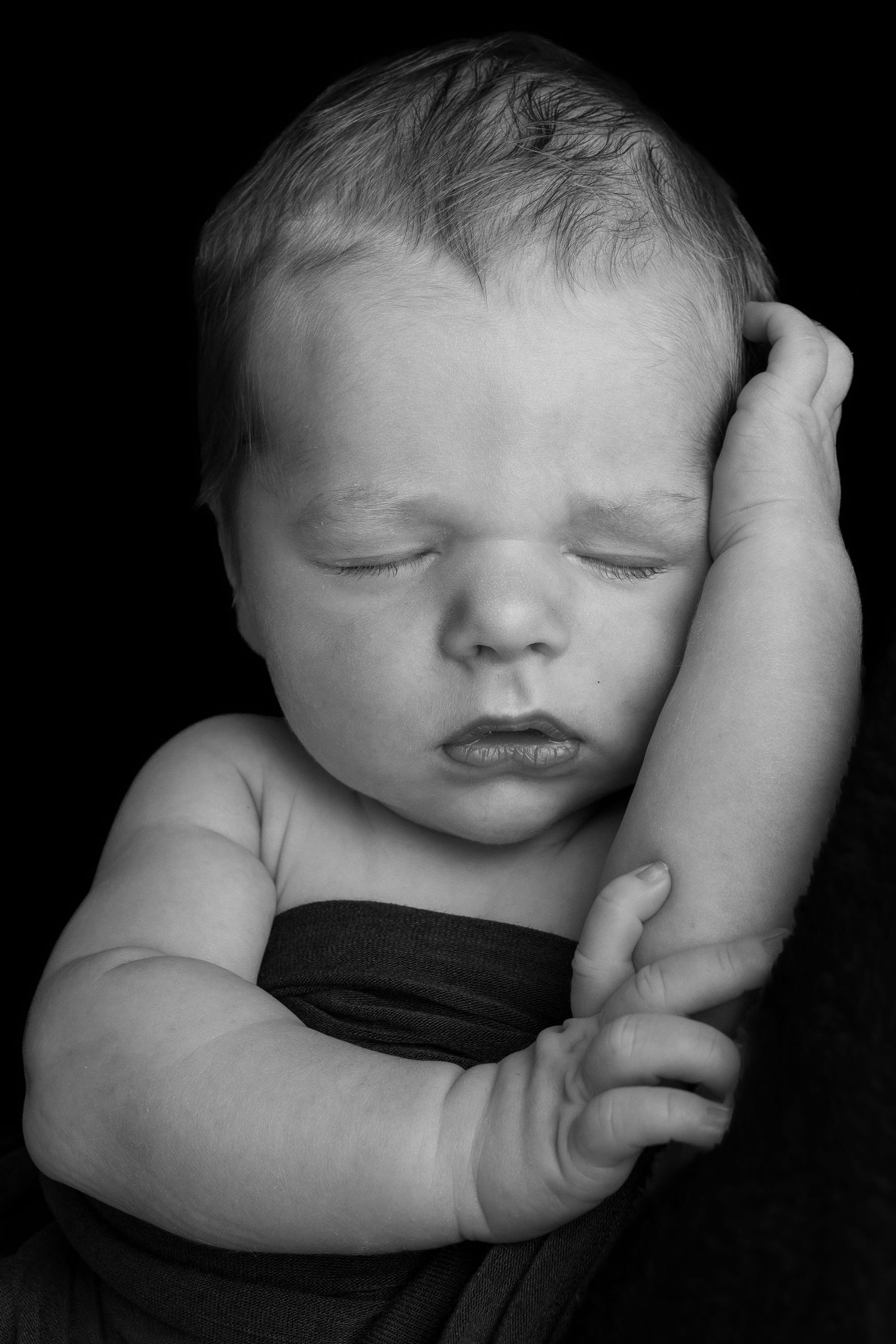 Black and white portrait of a sleeping newborn baby resting head on hand against dark background.