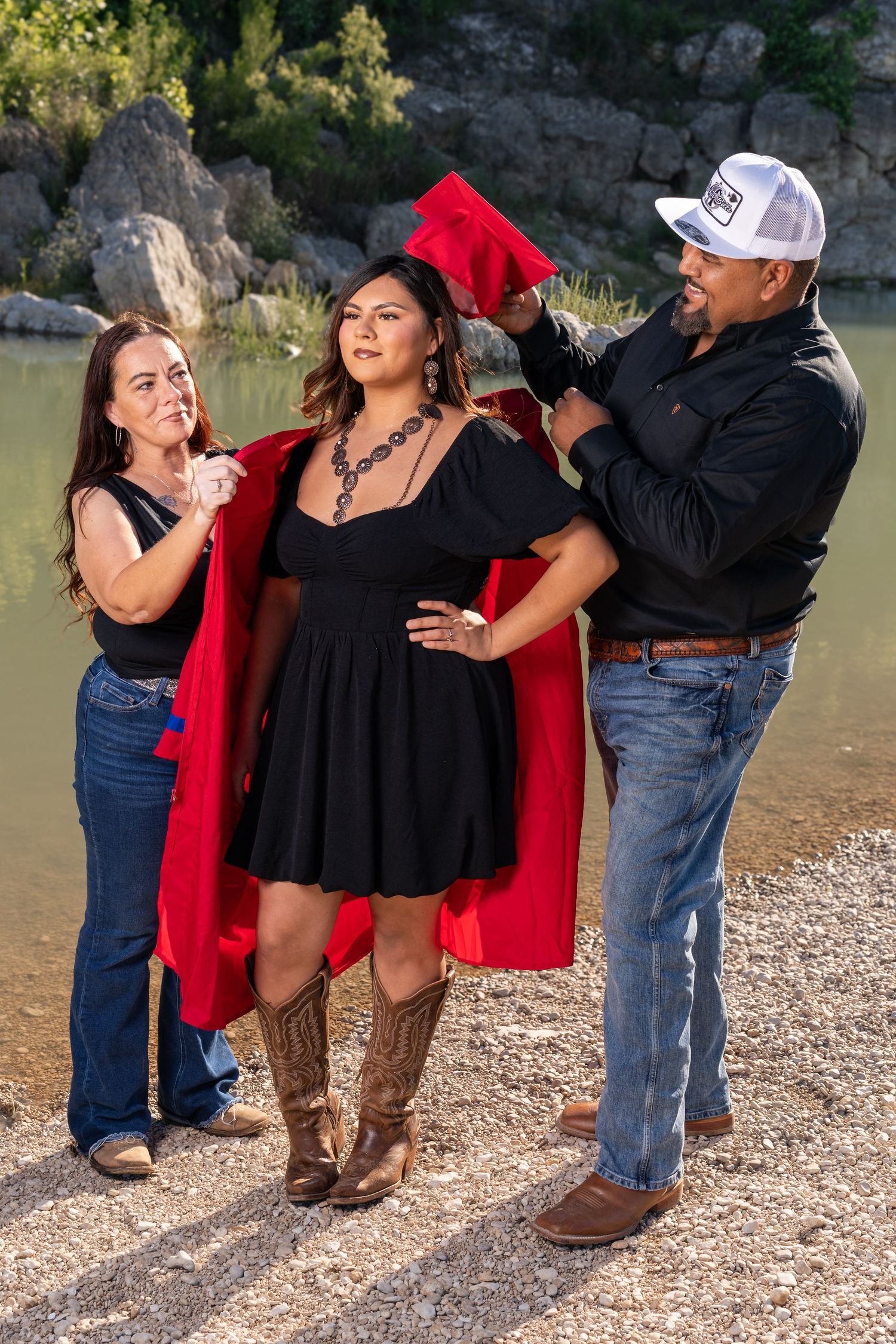 Parents help their daughter put on a red graduation cap and gown outdoors near a rocky river.