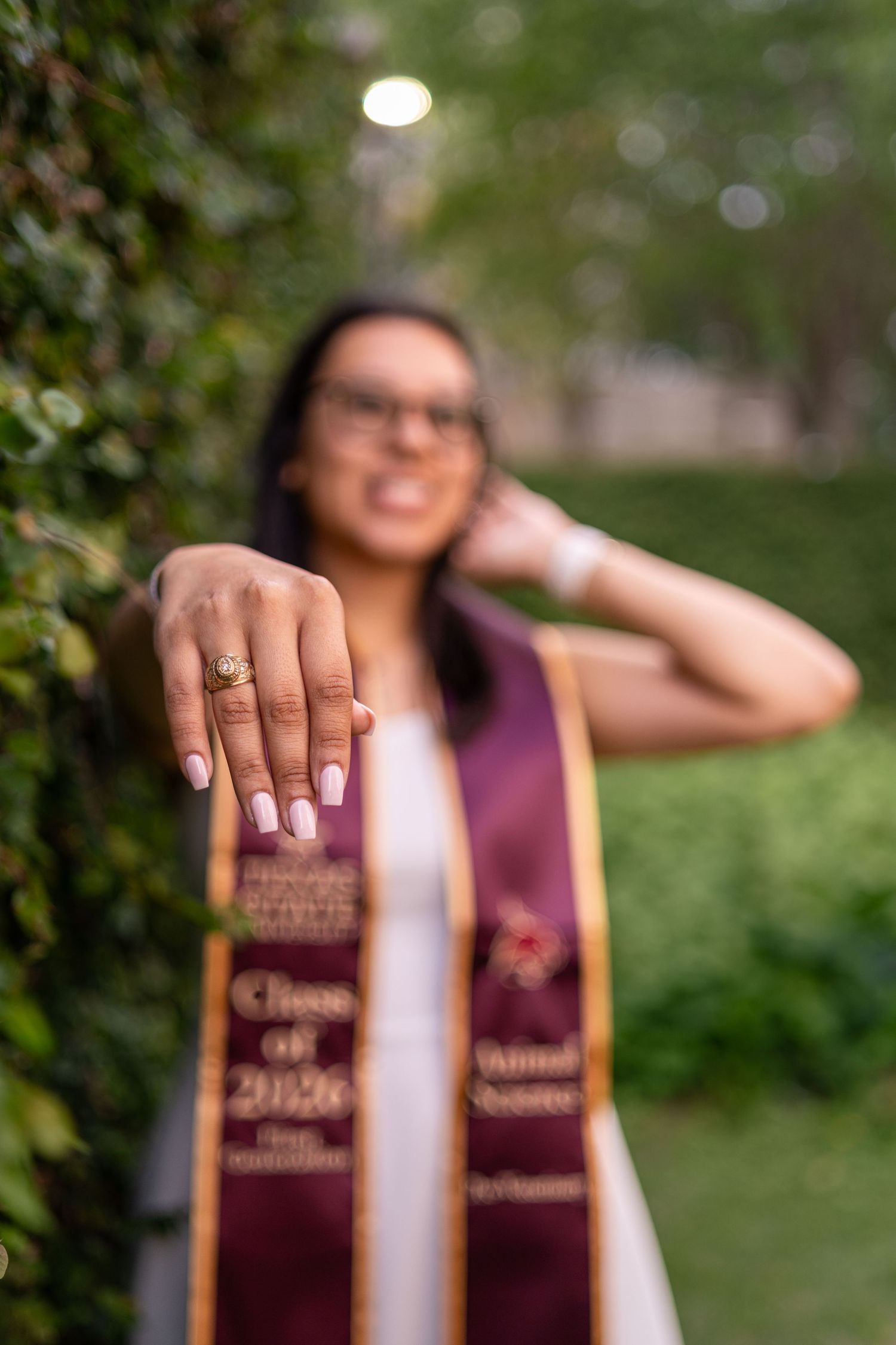Graduate showing off class ring wearing maroon Class of 2026 honor stole outdoors among green foliage.