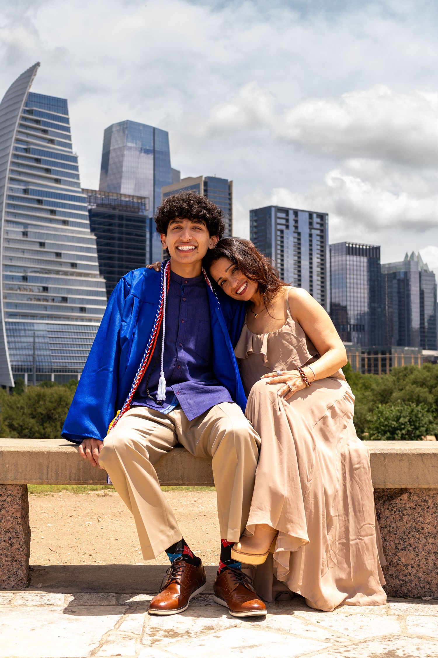 A smiling graduate in a blue cap and gown sits with a woman in a beige dress against a city skyline backdrop.