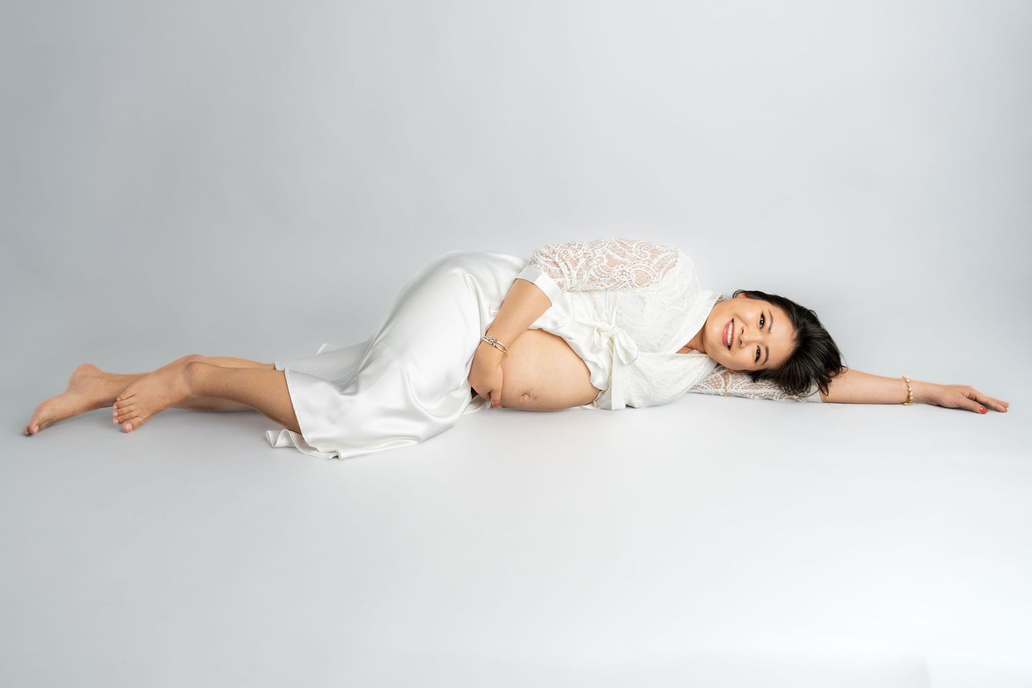 A pregnant woman in a white dress lying gracefully on a white studio floor with one arm extended.