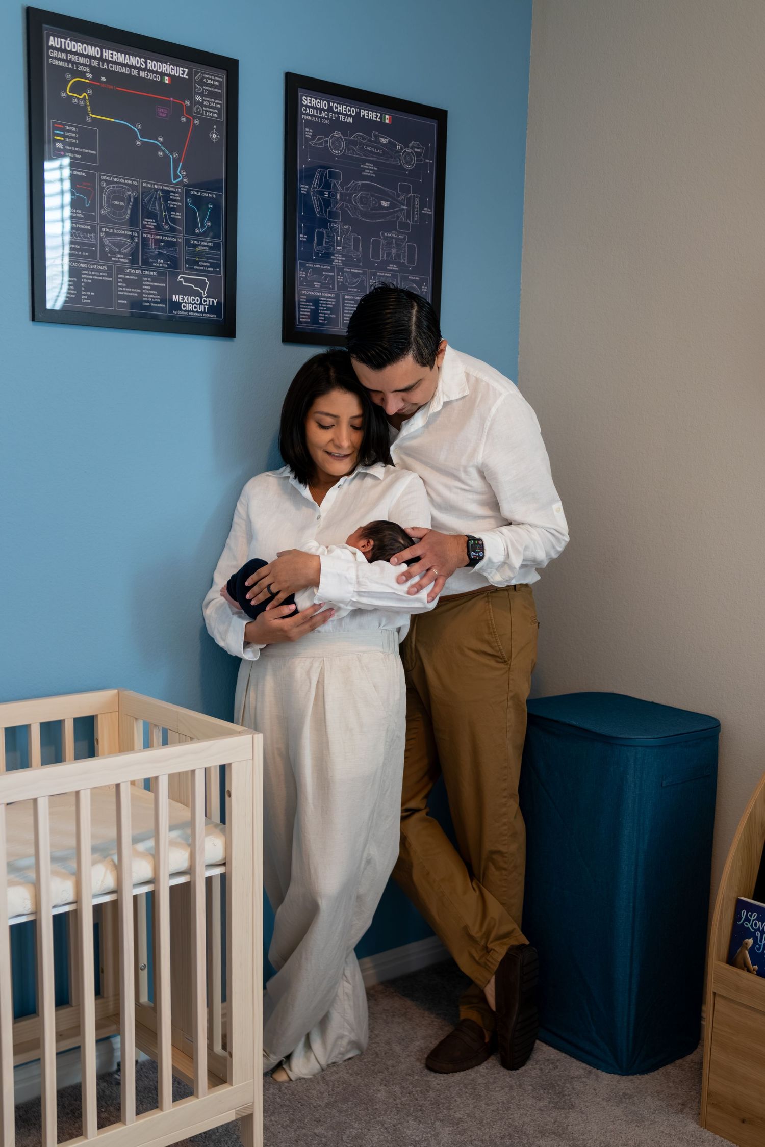 New parents cradle their newborn baby in a blue nursery, standing beside a white crib with framed prints on the wall.