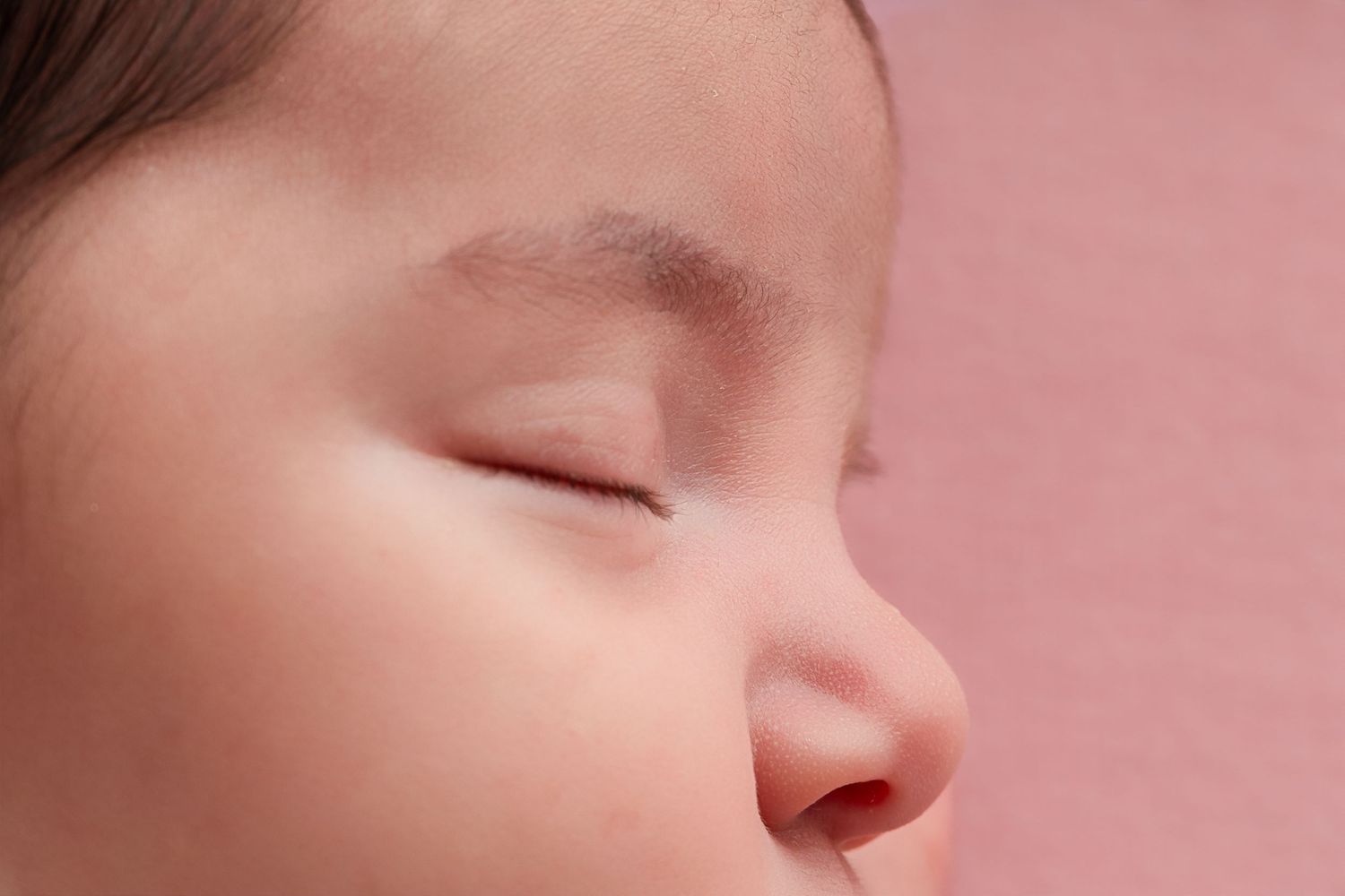 Close-up side profile of a sleeping baby's face with closed eyes and soft skin against a pink background.