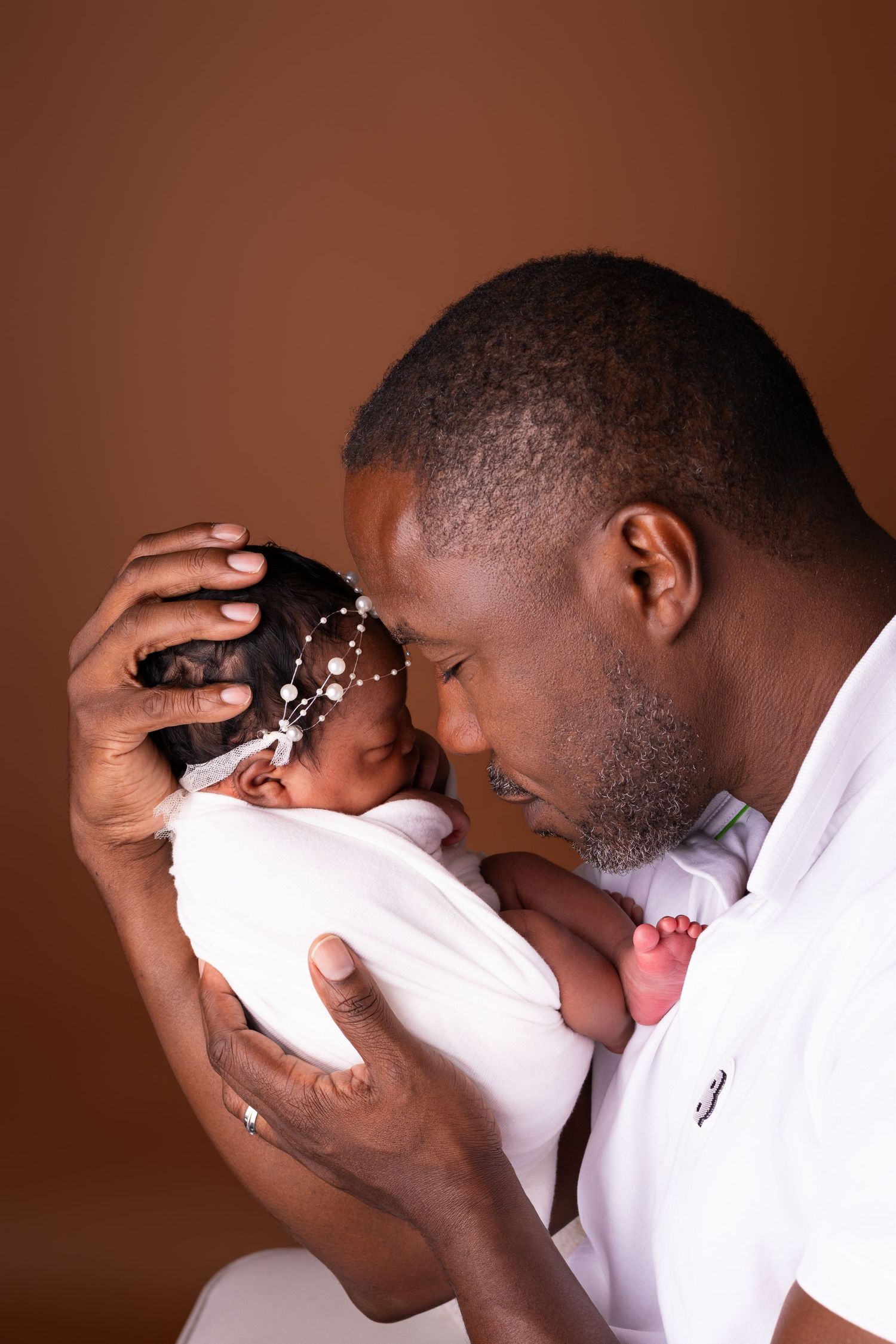 A father in a white polo gently touches foreheads with his sleeping newborn, who wears a pearl headband, in a warm newborn portrait.