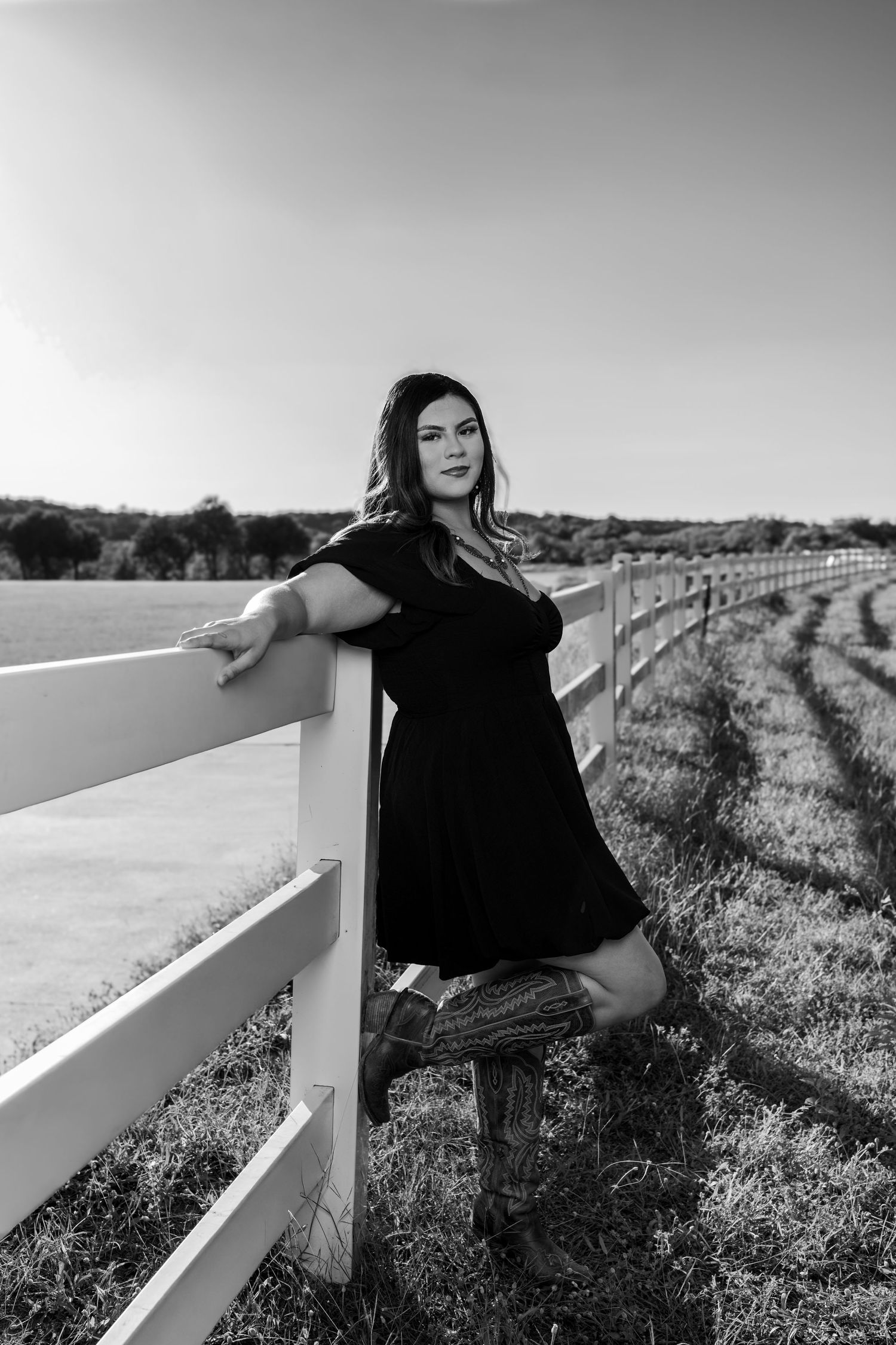 Woman in black dress and cowboy boots leaning on white fence in rural field, black and white photo.