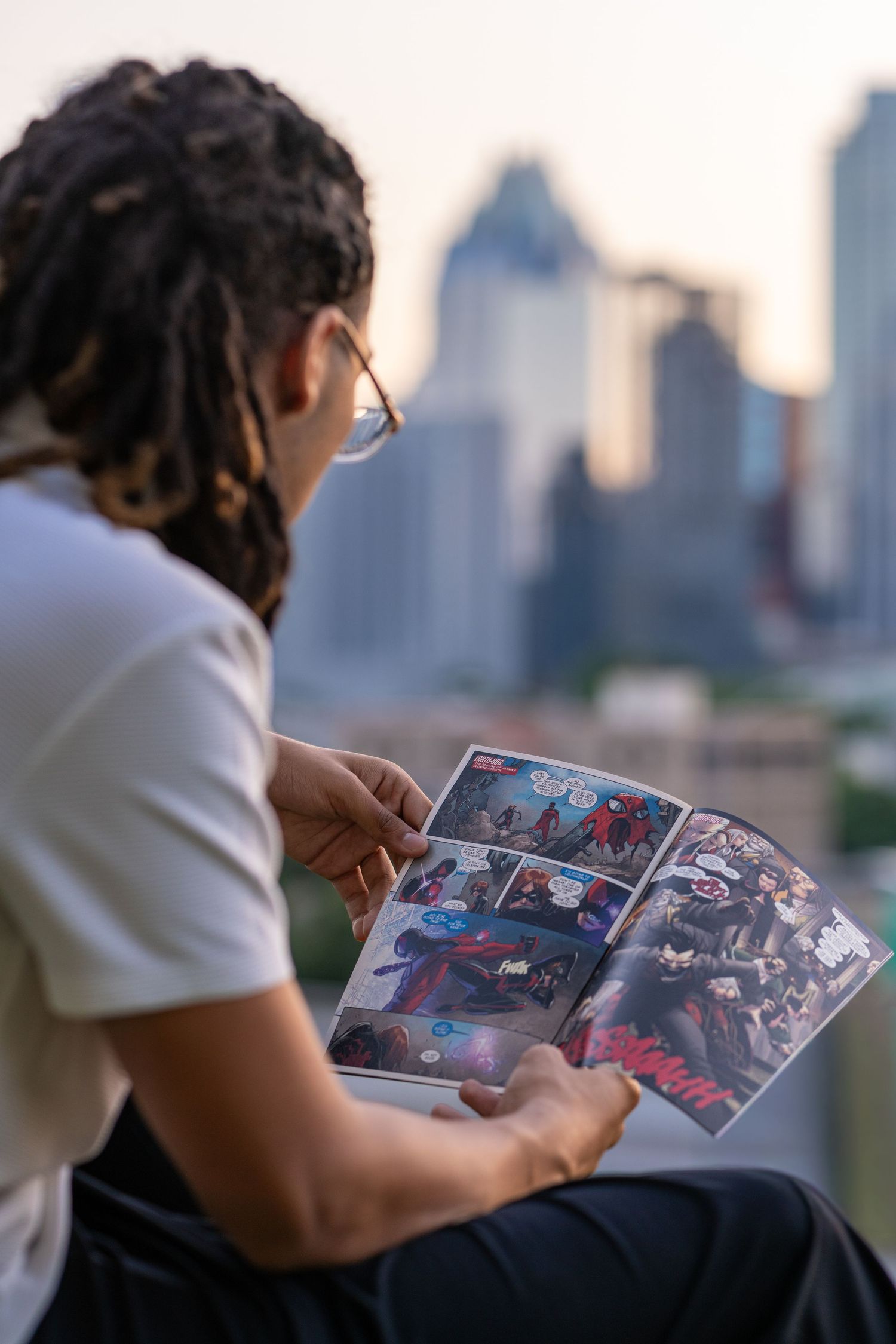 Person with dreadlocks reading a comic book on a rooftop with a city skyline at sunset.
