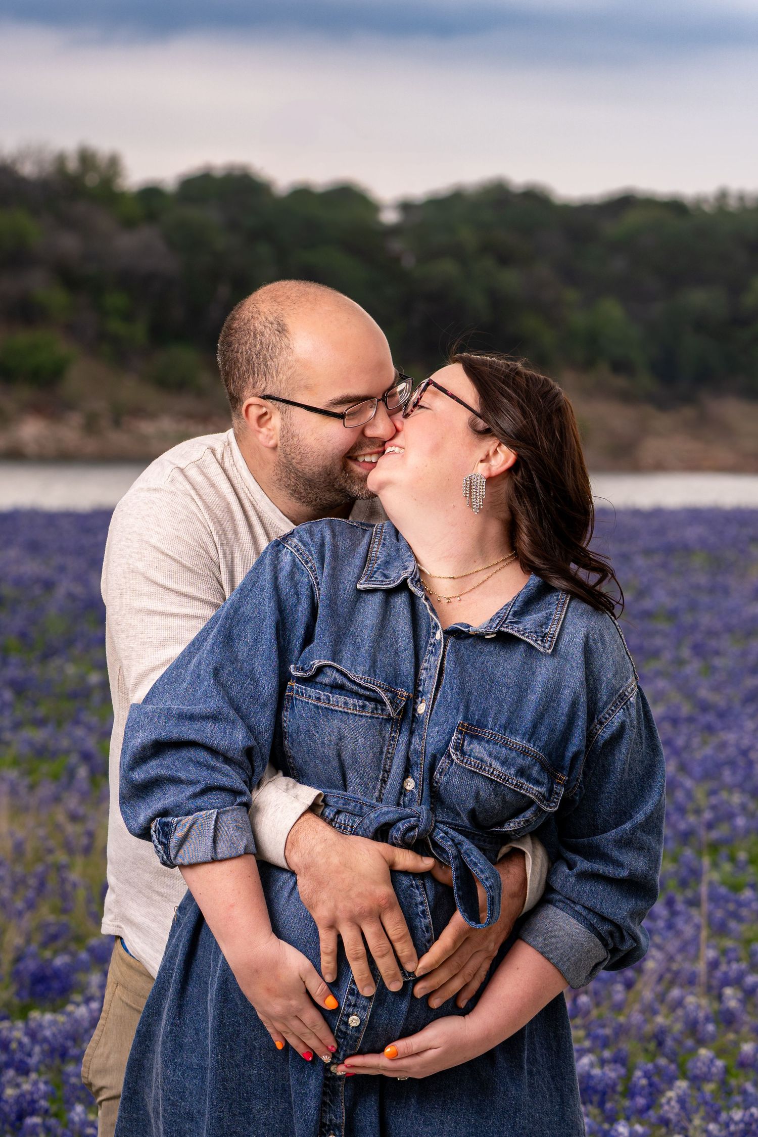 Couple sharing a tender kiss in a Texas bluebonnet field, man embracing pregnant woman in denim dress.
