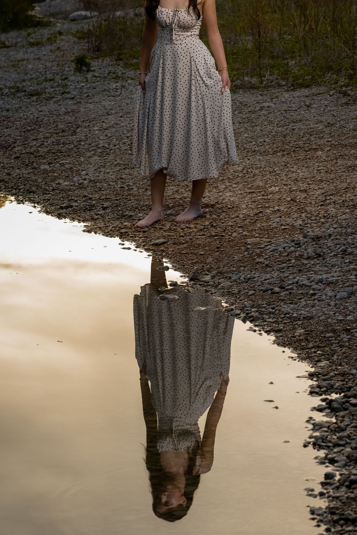 Woman in floral midi dress standing barefoot by a puddle reflecting her image on a rocky shore.