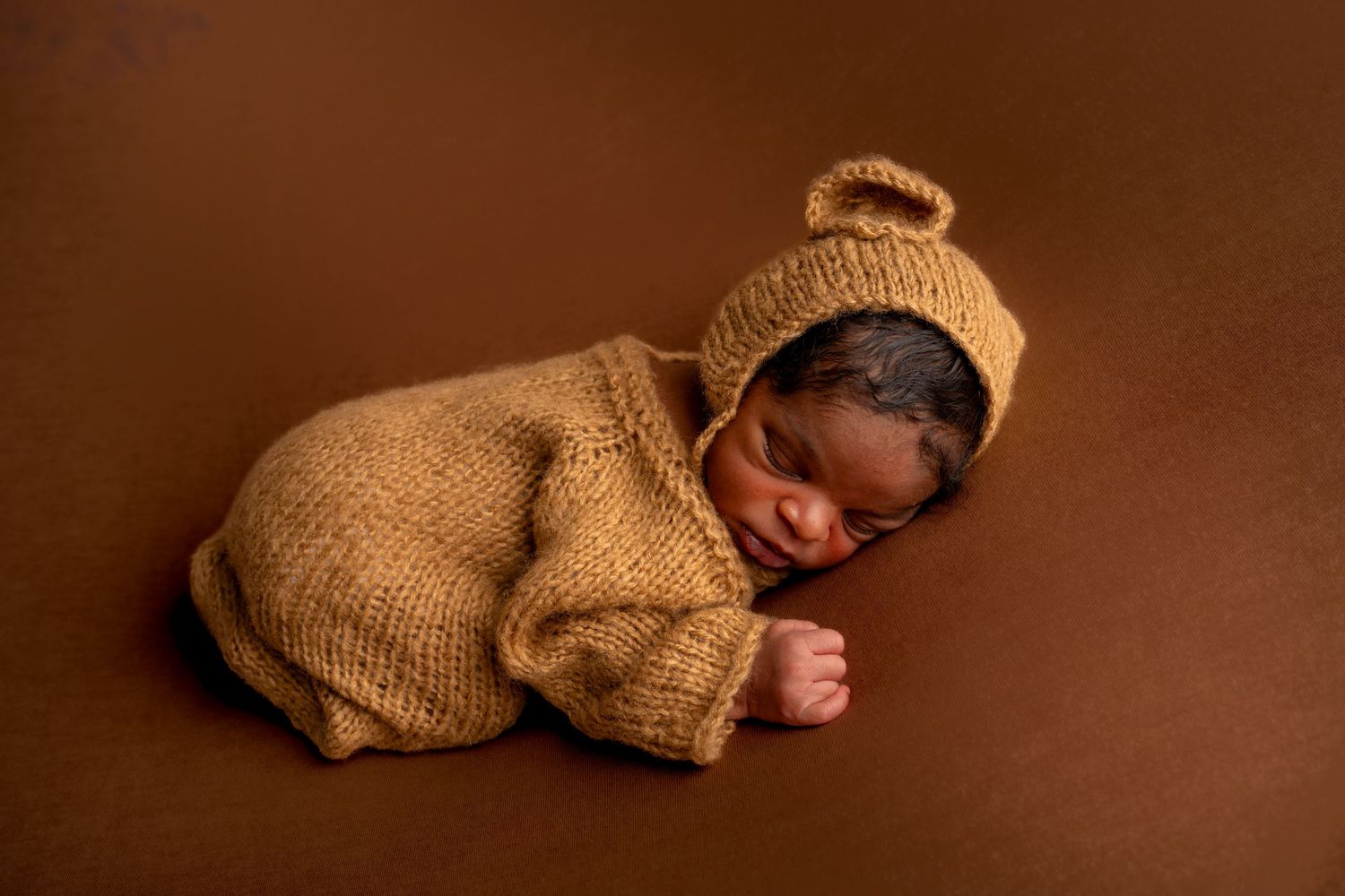 Newborn baby sleeping in brown knit bear romper and hat on brown background.