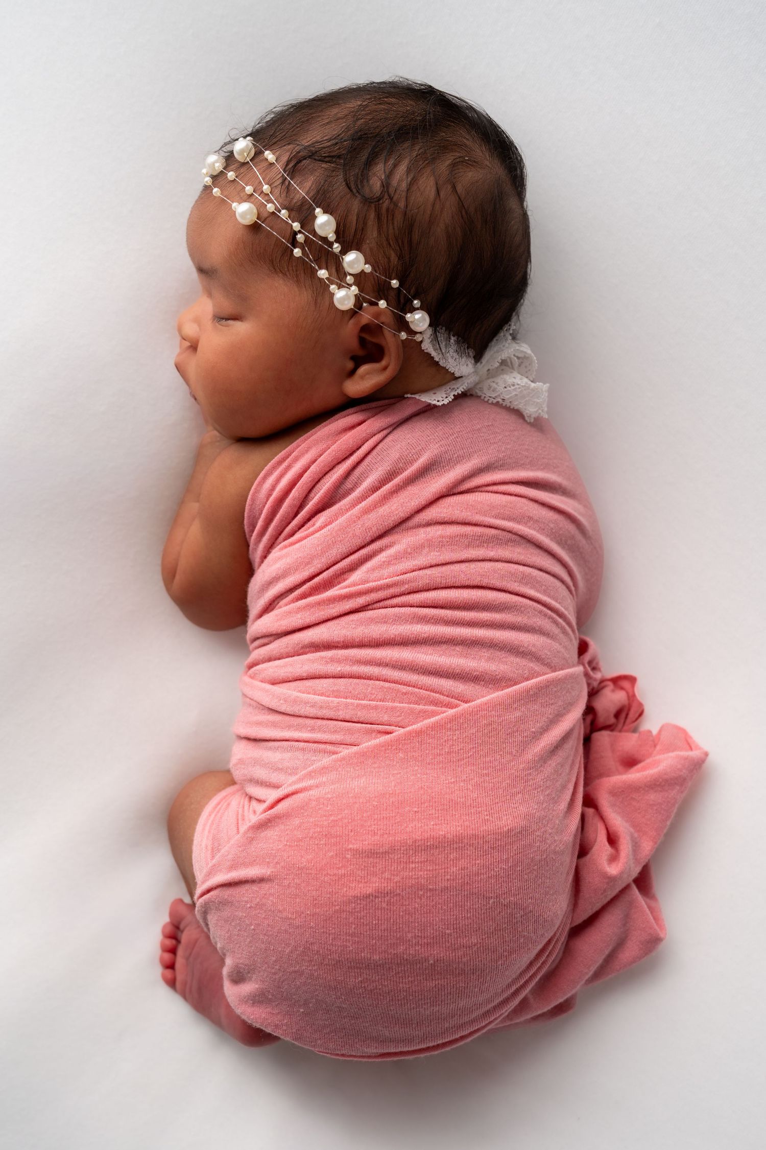Sleeping newborn baby girl swaddled in pink wrap wearing a delicate pearl headband on white background.